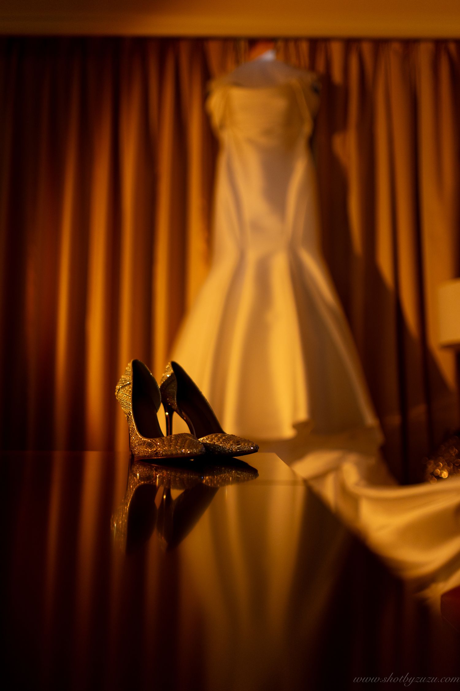 Wedding dress silhouetted against orange curtains with shoes reflected on glossy surface.