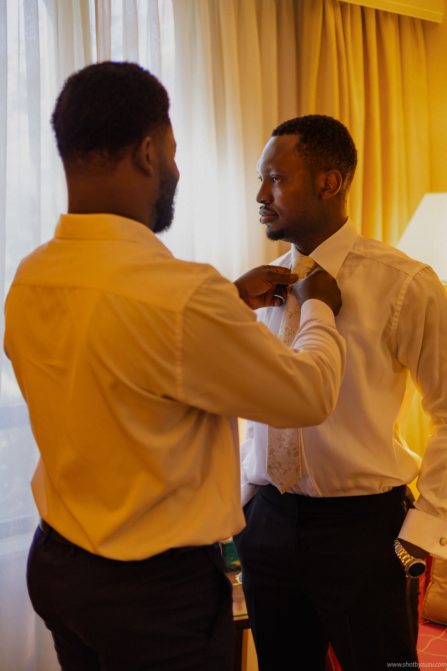 Wedding preparation scene with formal attire adjustment in warm yellow-lit room.