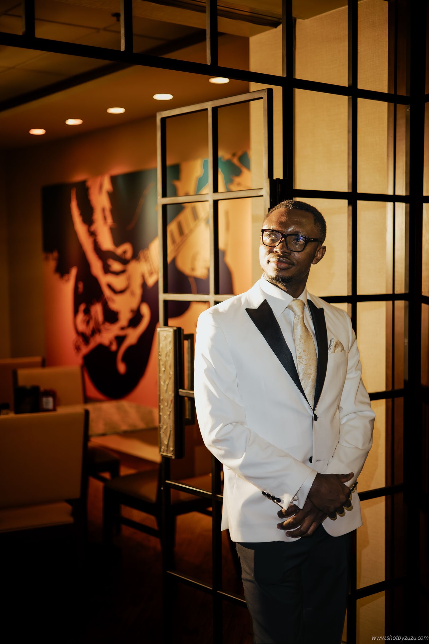 Stylish groom in white dinner jacket and glasses poses against modern interior with artistic backdrop.