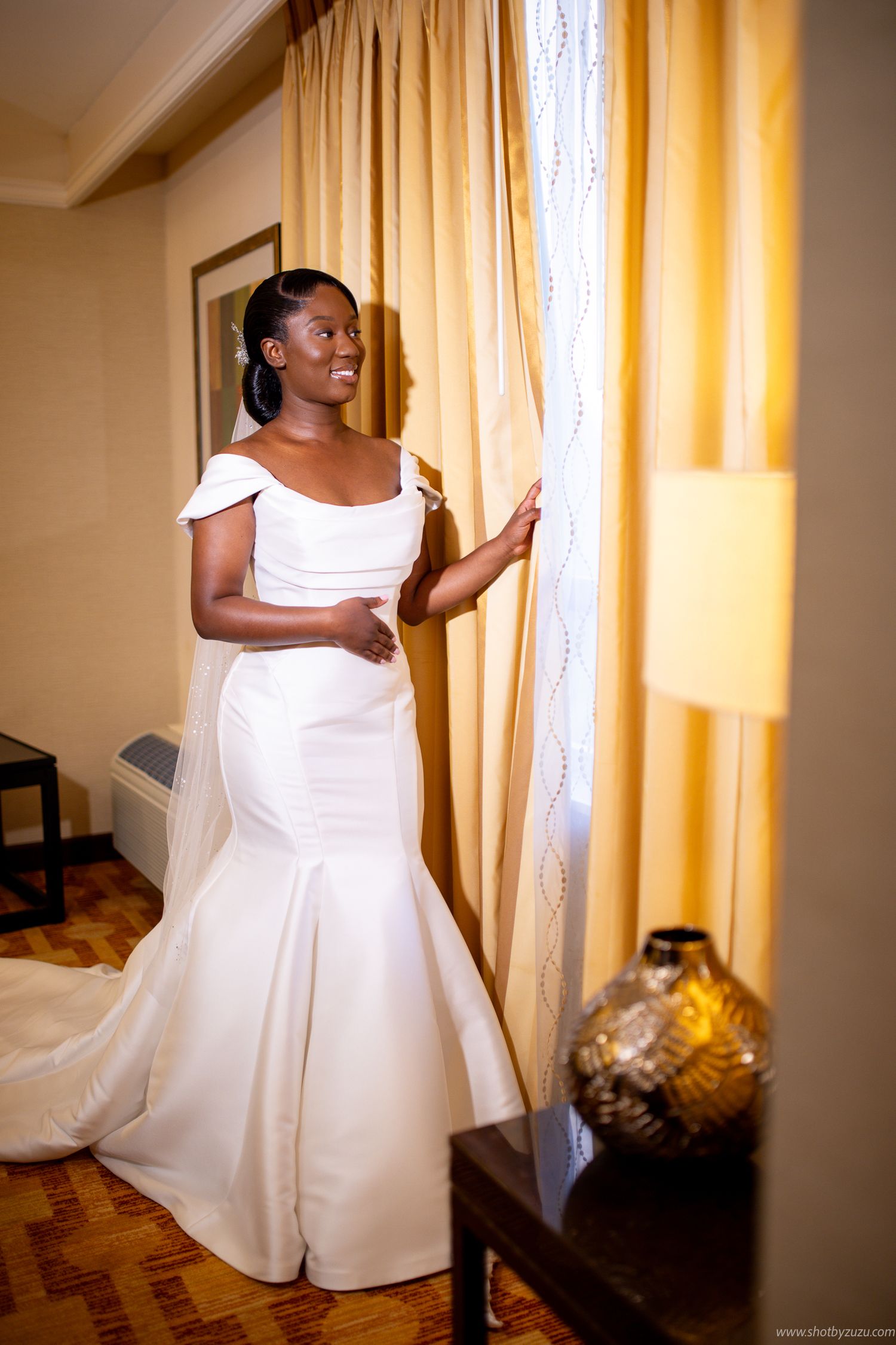 Full-length shot of bride in fitted white gown with cap sleeves standing by window.