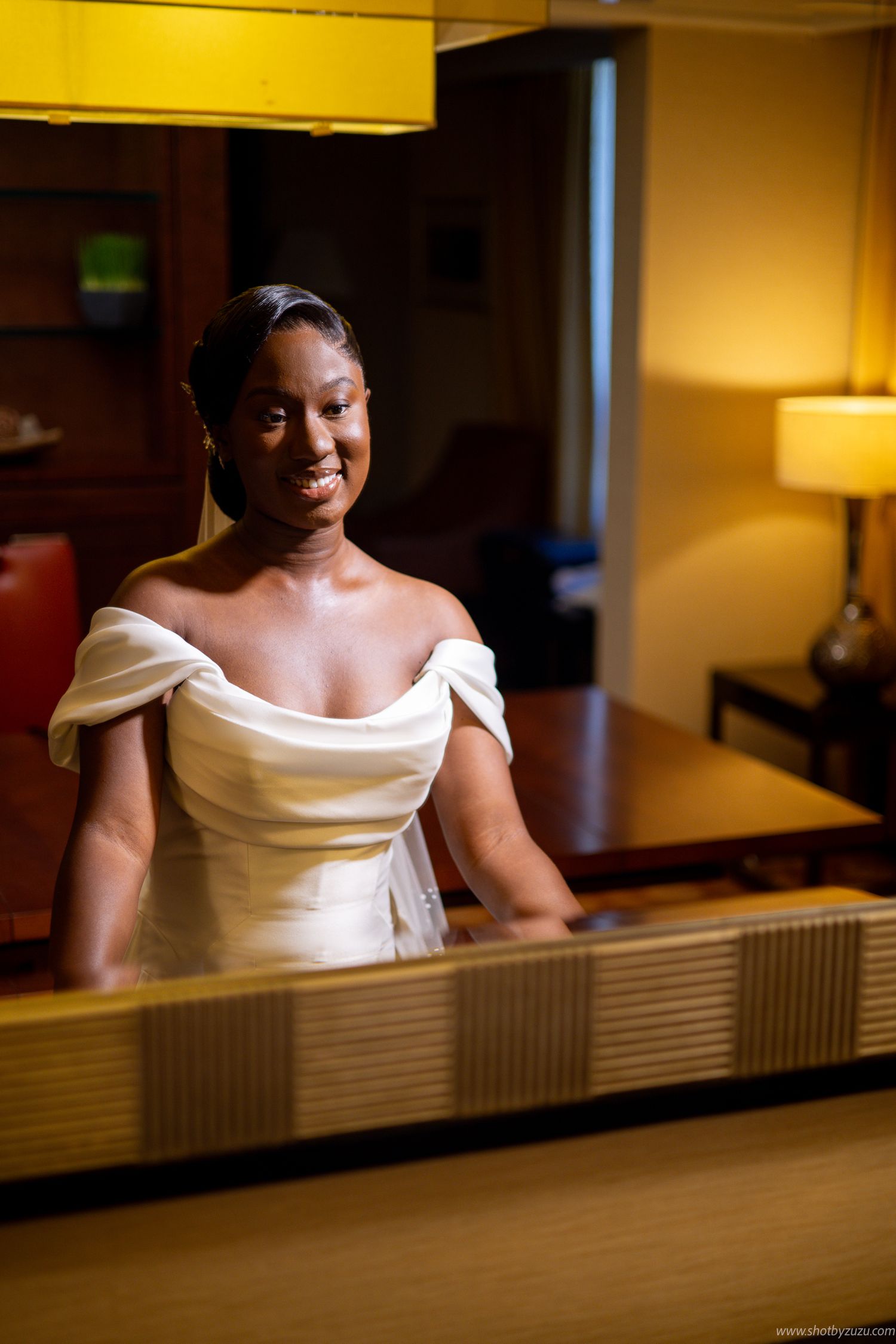 A bride in a white off-shoulder wedding dress smiles warmly while standing in a warmly lit room.