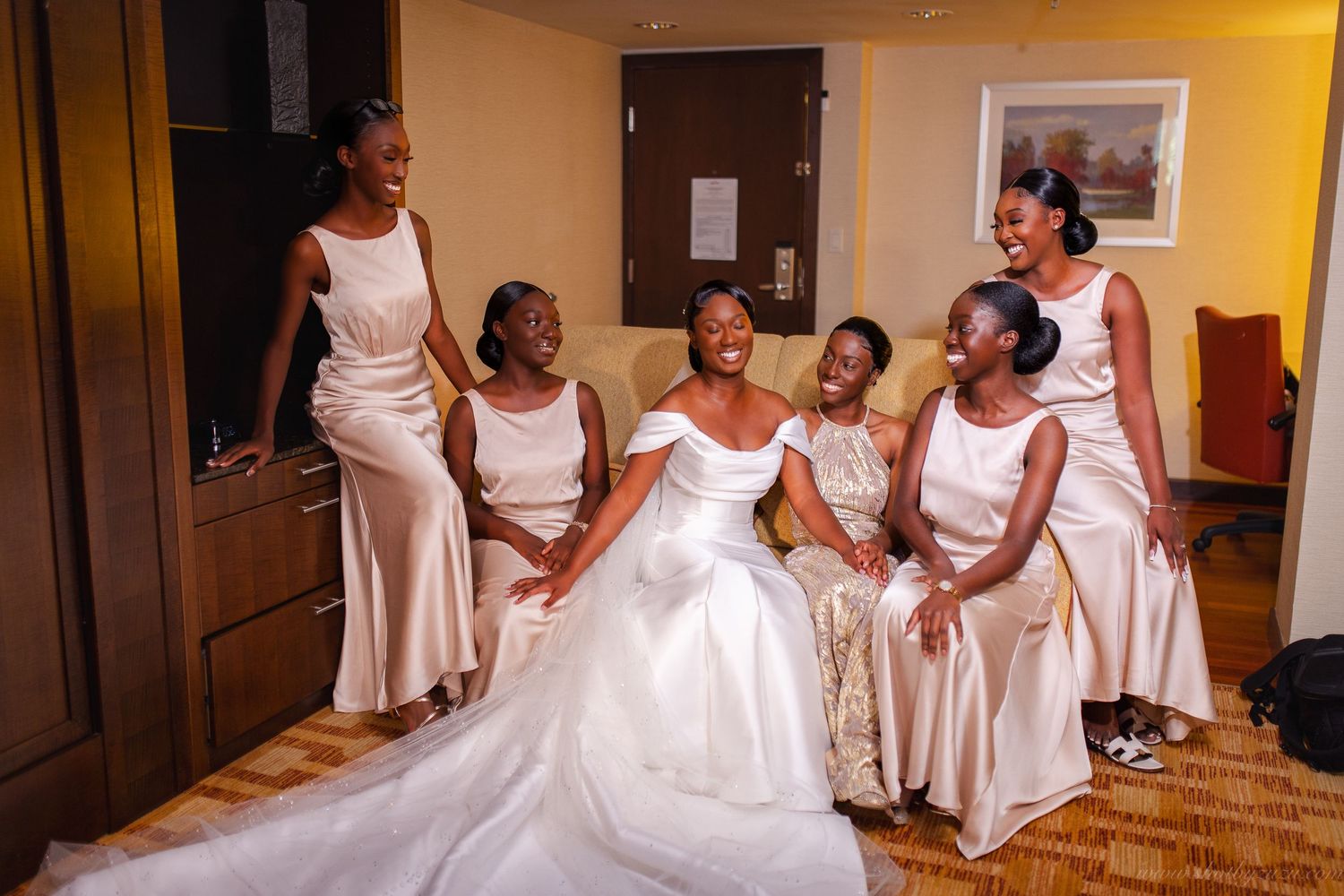 Wedding party in matching cream gowns surrounds bride in traditional white dress in hotel room.