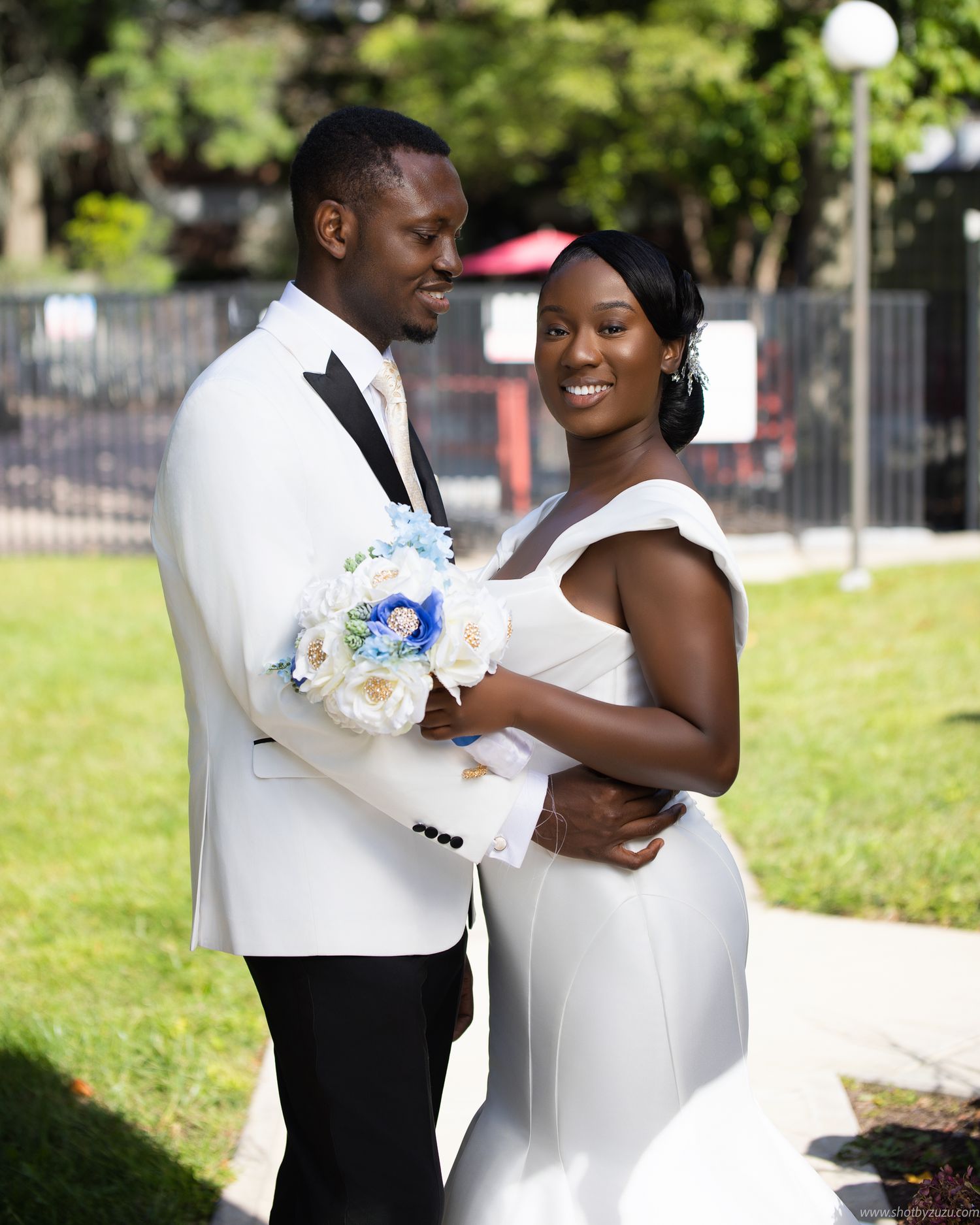A bright outdoor wedding portrait with white floral bouquet and classic white tuxedo against garden scenery.
