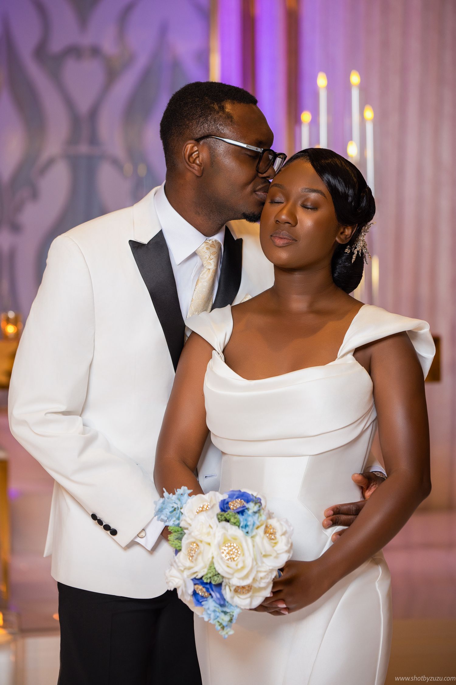 An intimate wedding portrait with a bouquet of white and blue flowers, captured in a pink-lit reception venue.