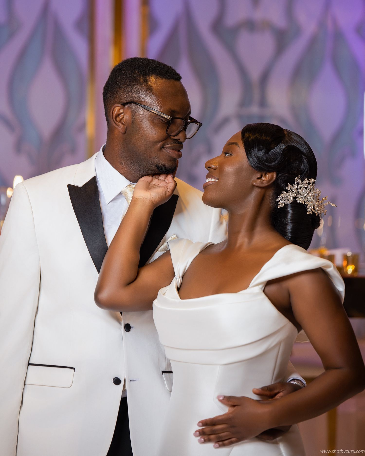 A joyful wedding couple shares a loving moment in front of a decorative backdrop with elegant lighting.