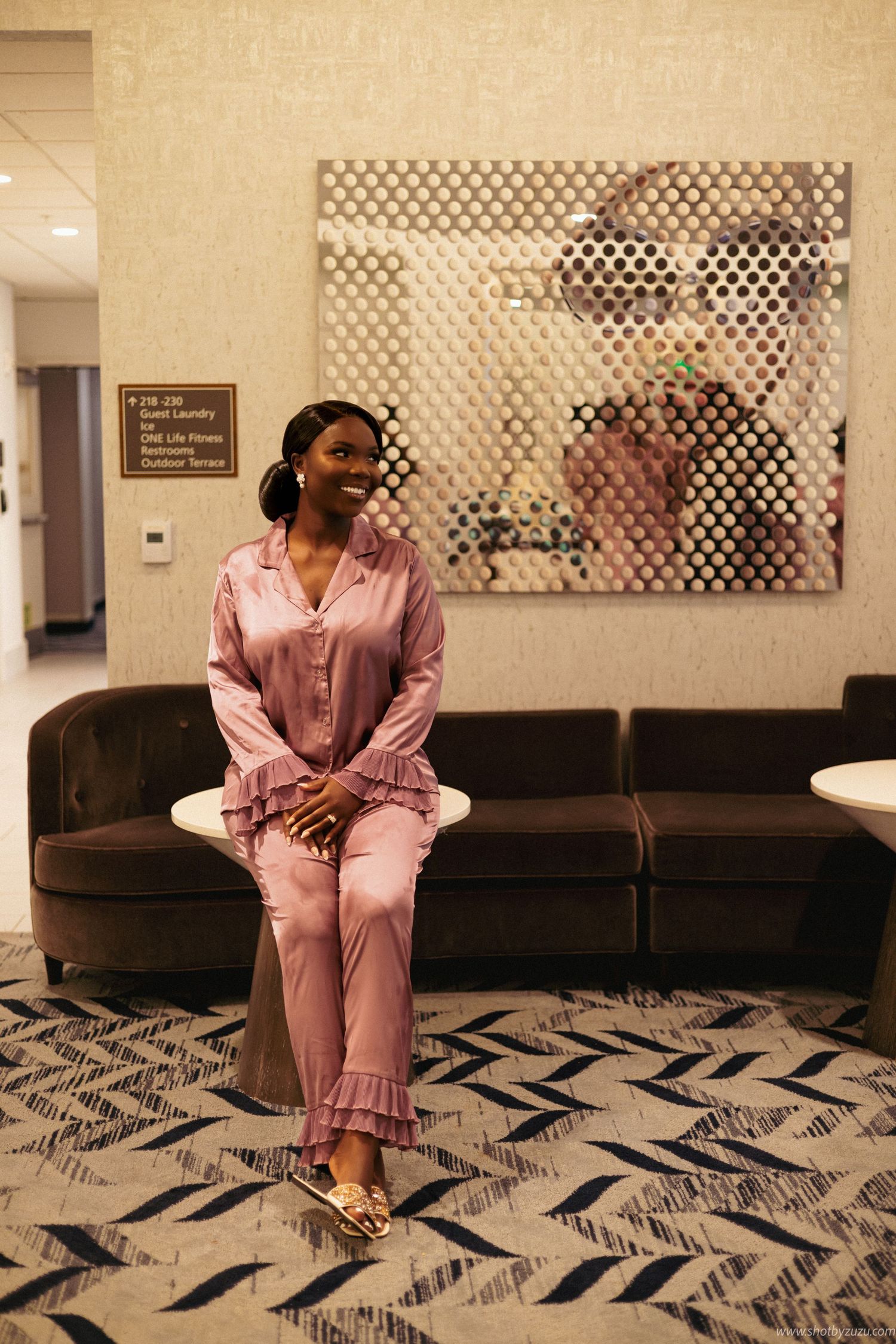 Woman in pink satin pajamas sitting on brown couch with geometric pattern carpet and wall art.