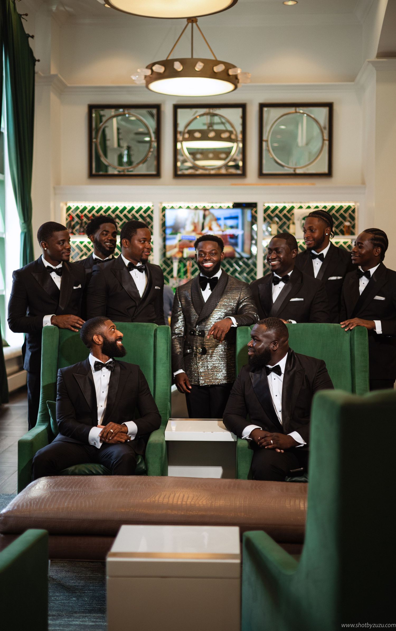 Groomsmen in black tuxedos gather in luxurious lounge with green velvet chairs and modern lighting.