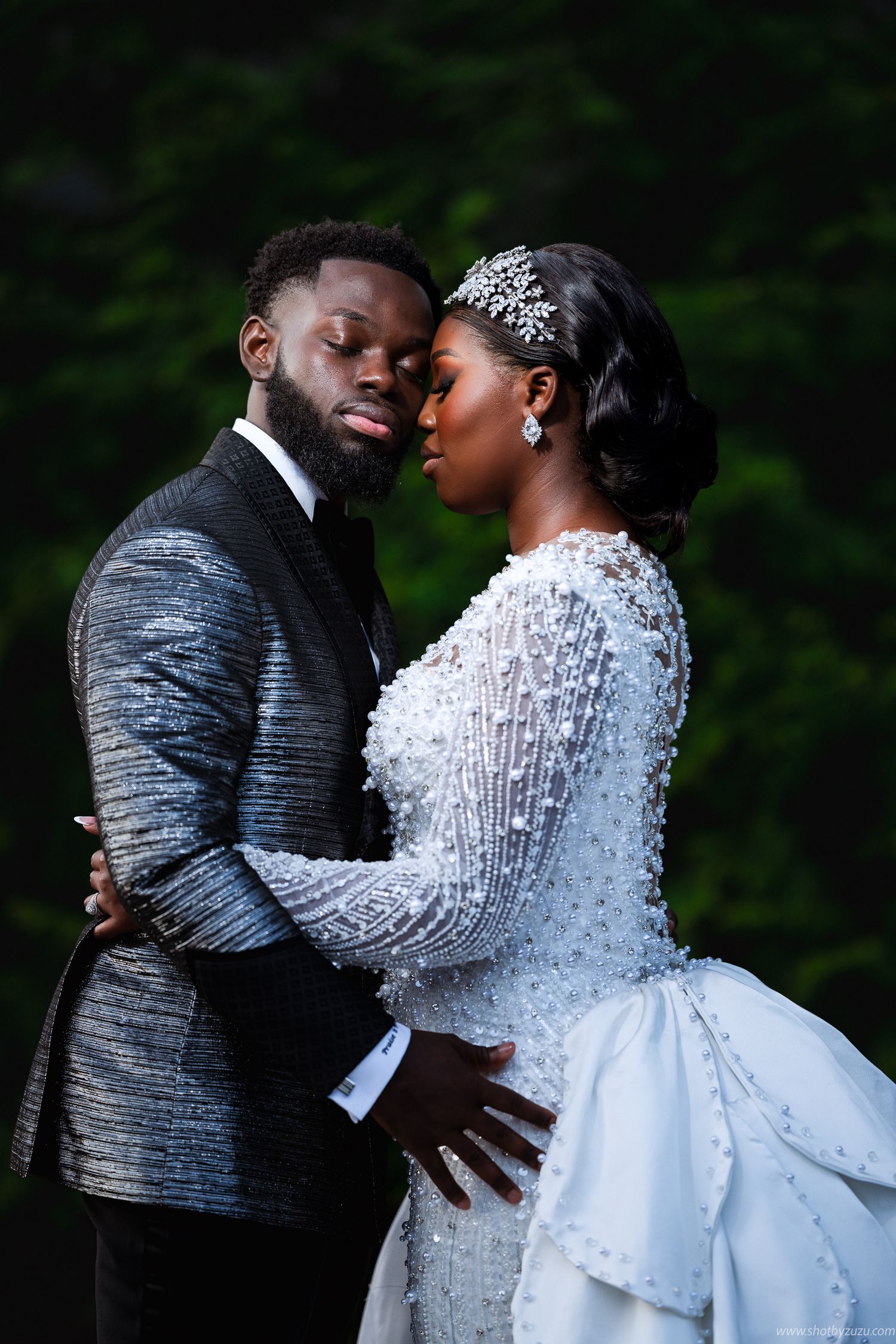 A romantic wedding portrait of a couple embracing outdoors, with the bride in a beaded white gown and sparkling tiara.