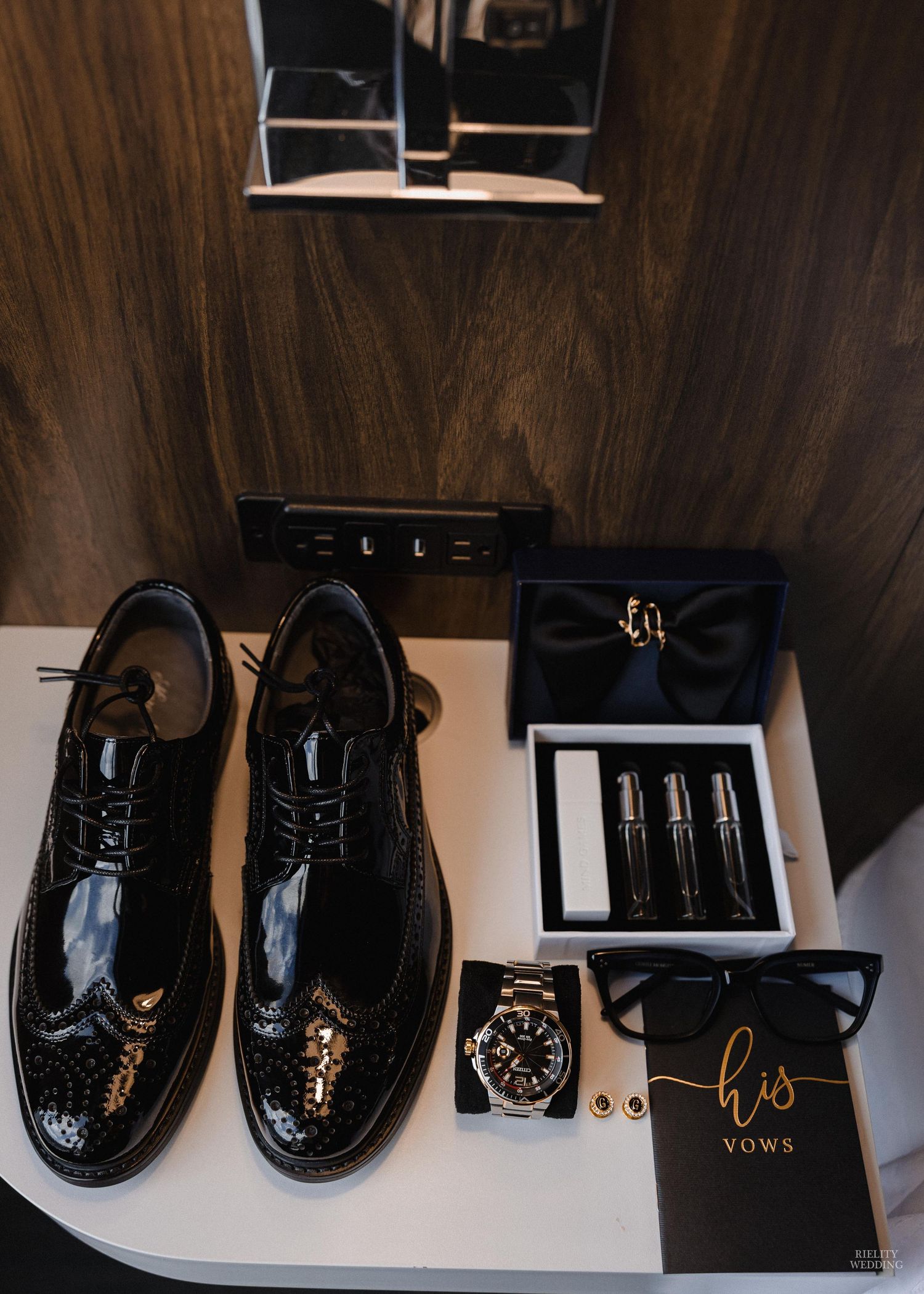 A row of formal black dress shoes and accessories displayed on a white surface against a dark wood wall.
