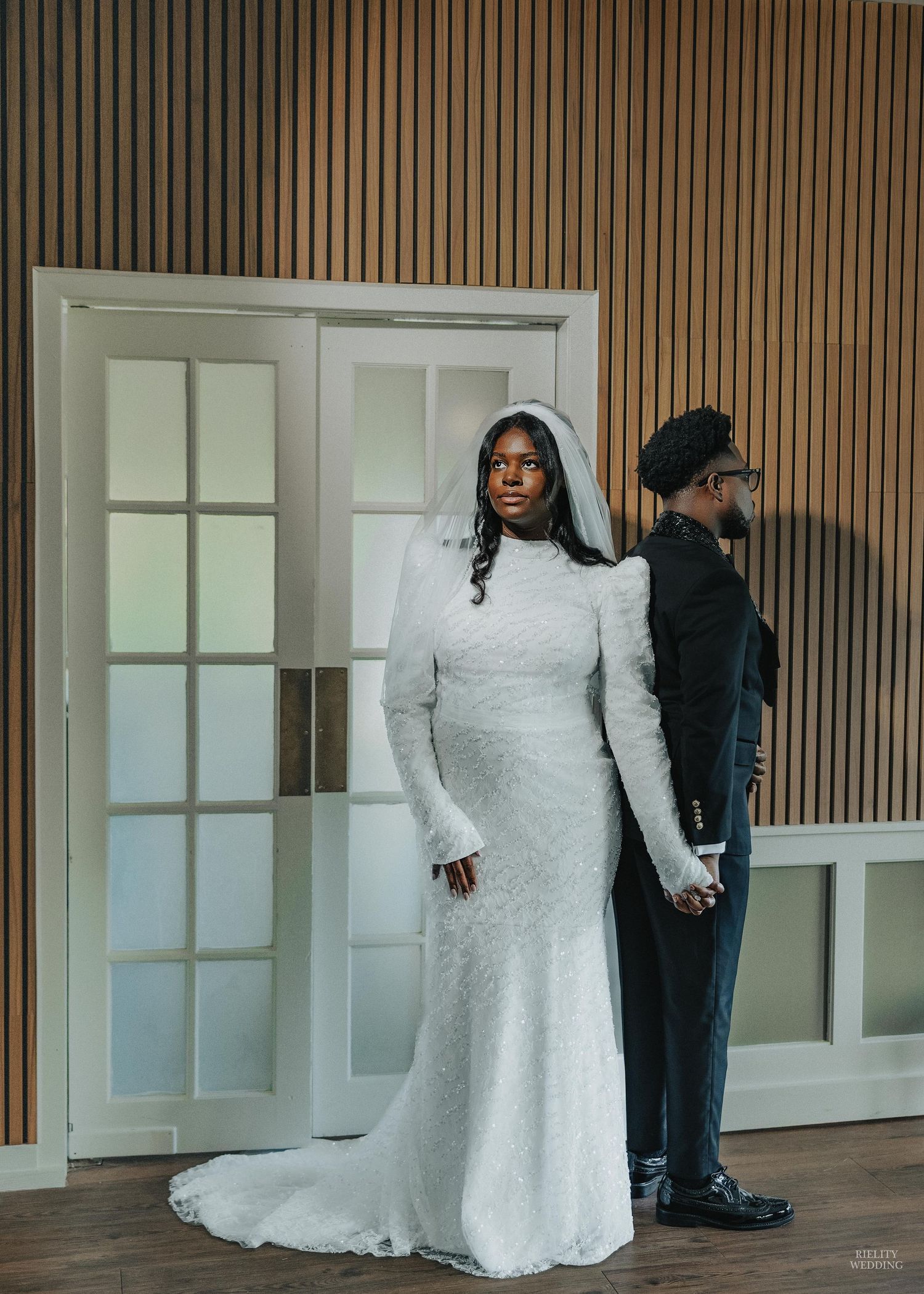 A white lace wedding dress displayed in a modern venue with wood paneled walls and glass doors.