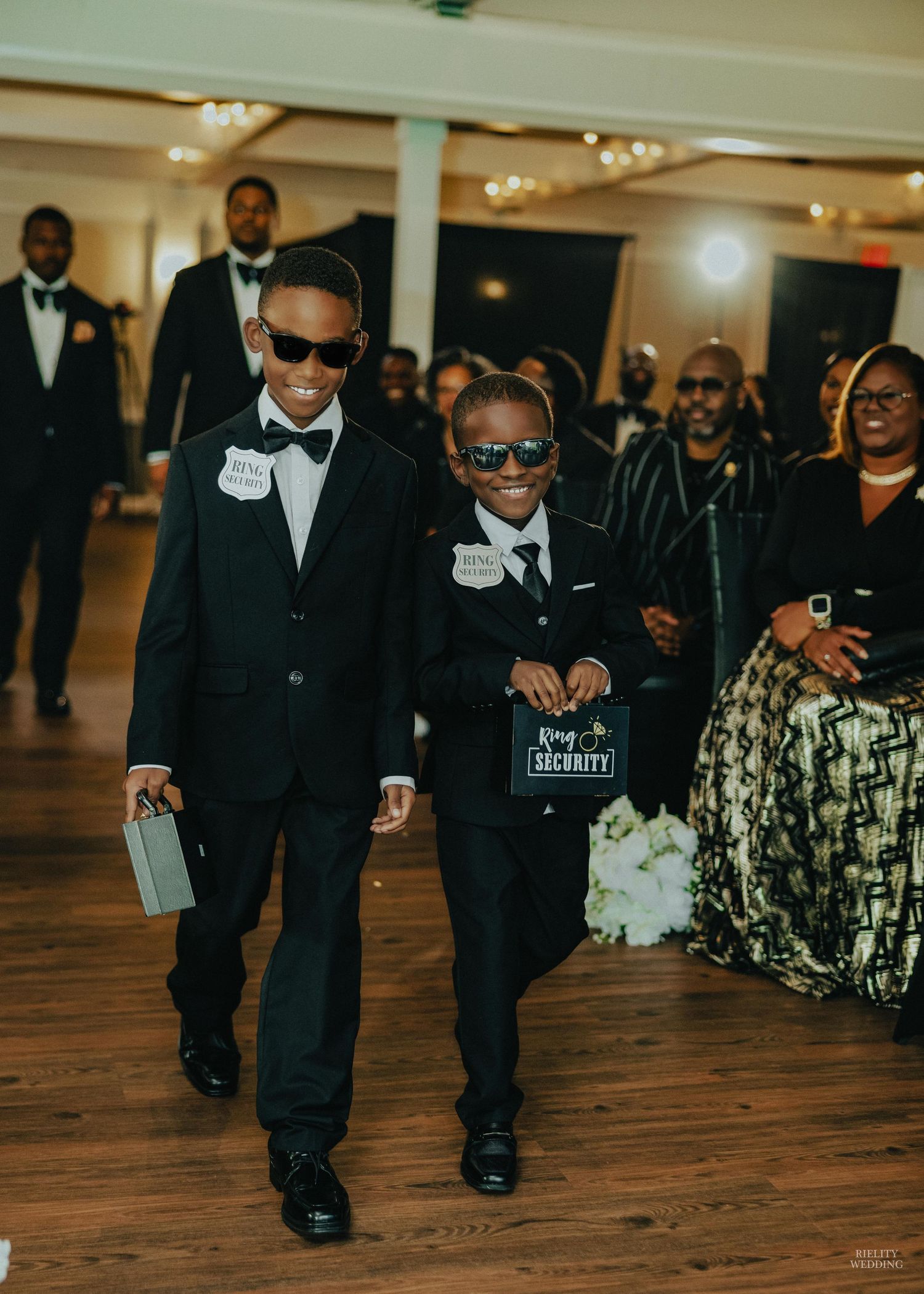 Ring bearers in black suits and sunglasses walk down a wooden floor aisle during a wedding ceremony.