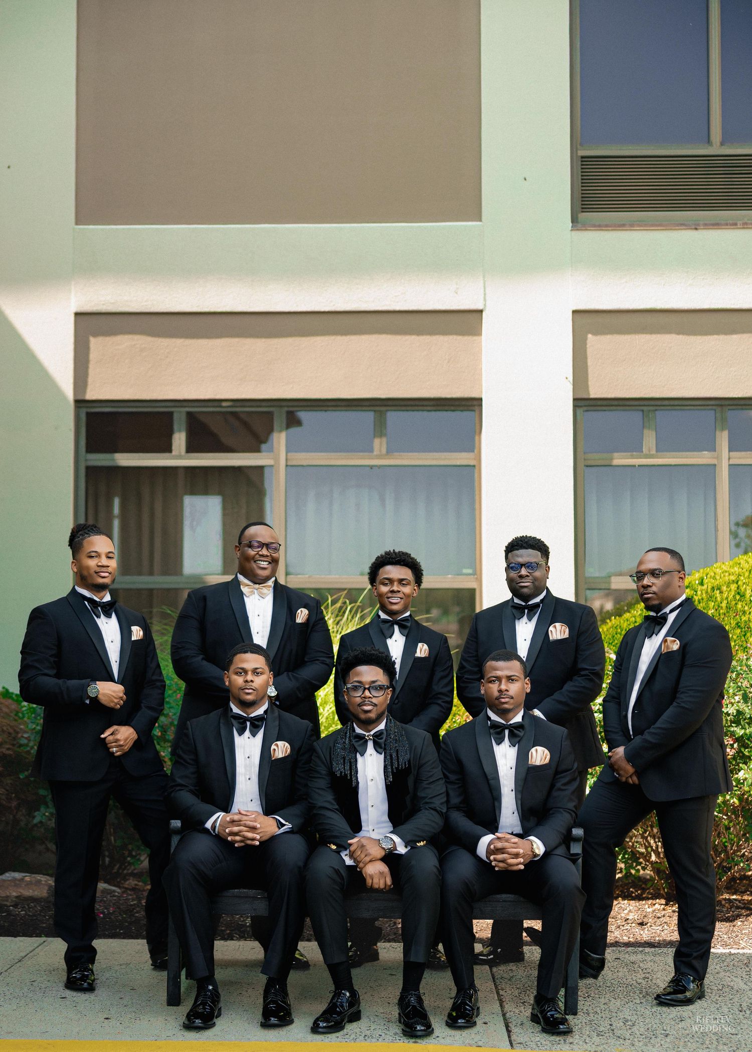 A panoramic photo of groomsmen in black tuxedos sitting on steps outside a building.