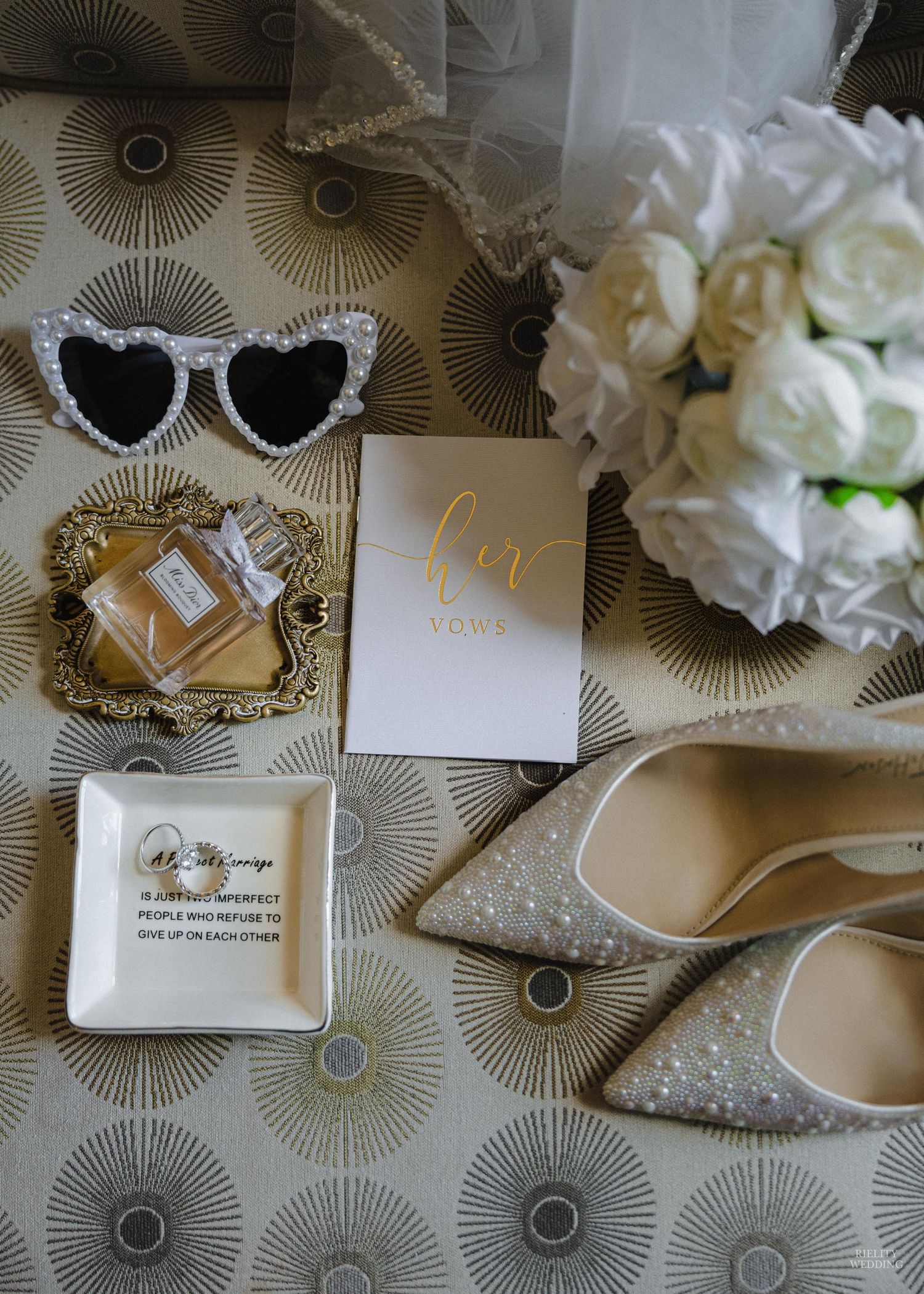 Elegant wedding accessories arranged on a patterned surface with white flowers, pearl details, and gold accents.
