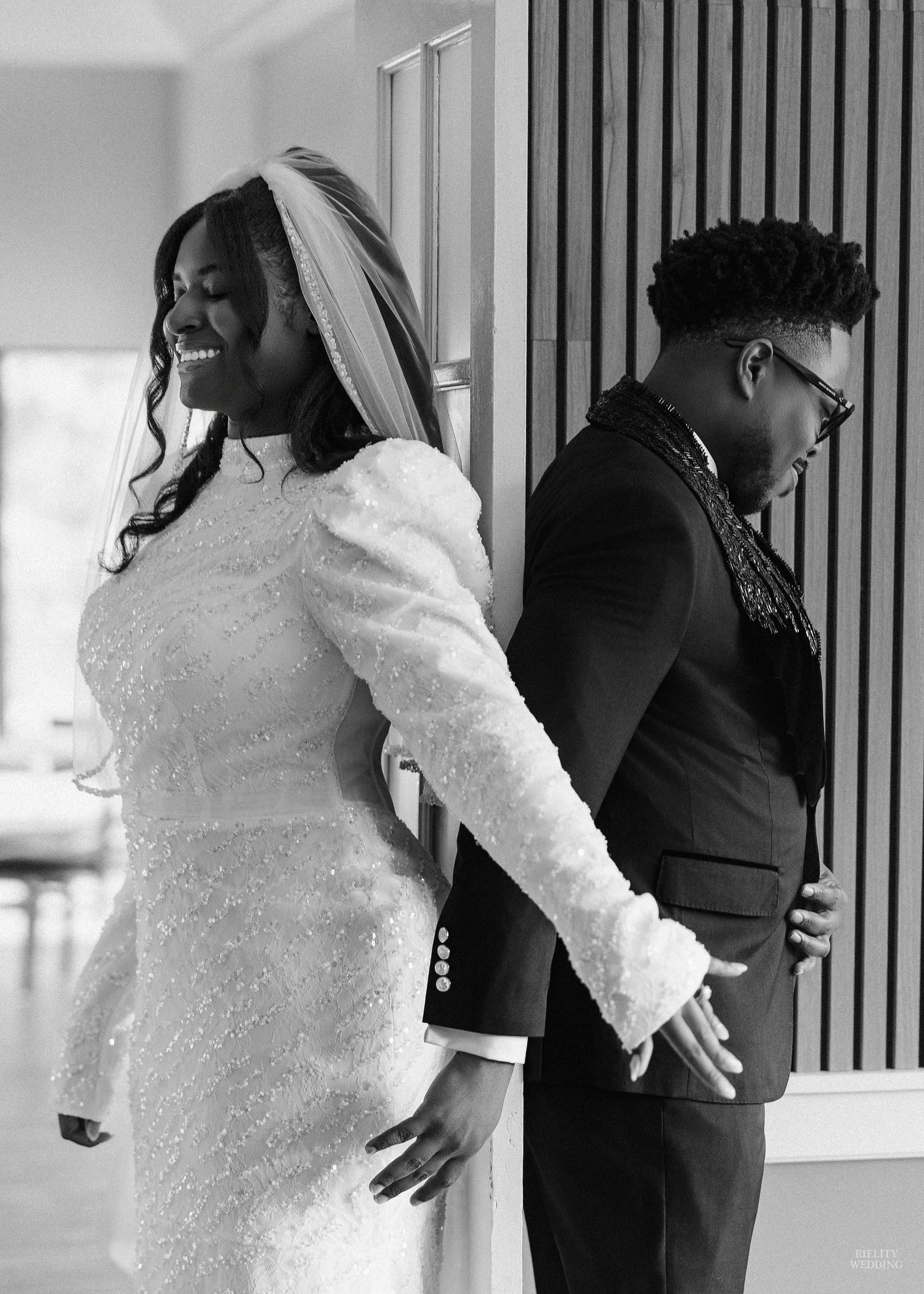 A bride and groom stand back-to-back during their wedding photoshoot, with the bride in white lace and groom in black suit.