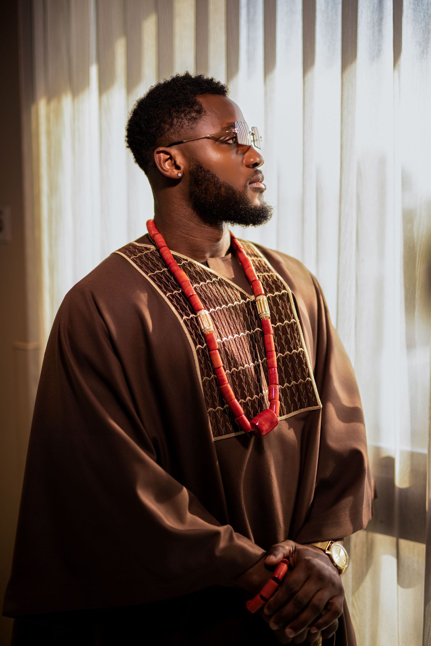 Portrait of person in traditional brown agbada with red beaded necklace against white curtain backdrop.