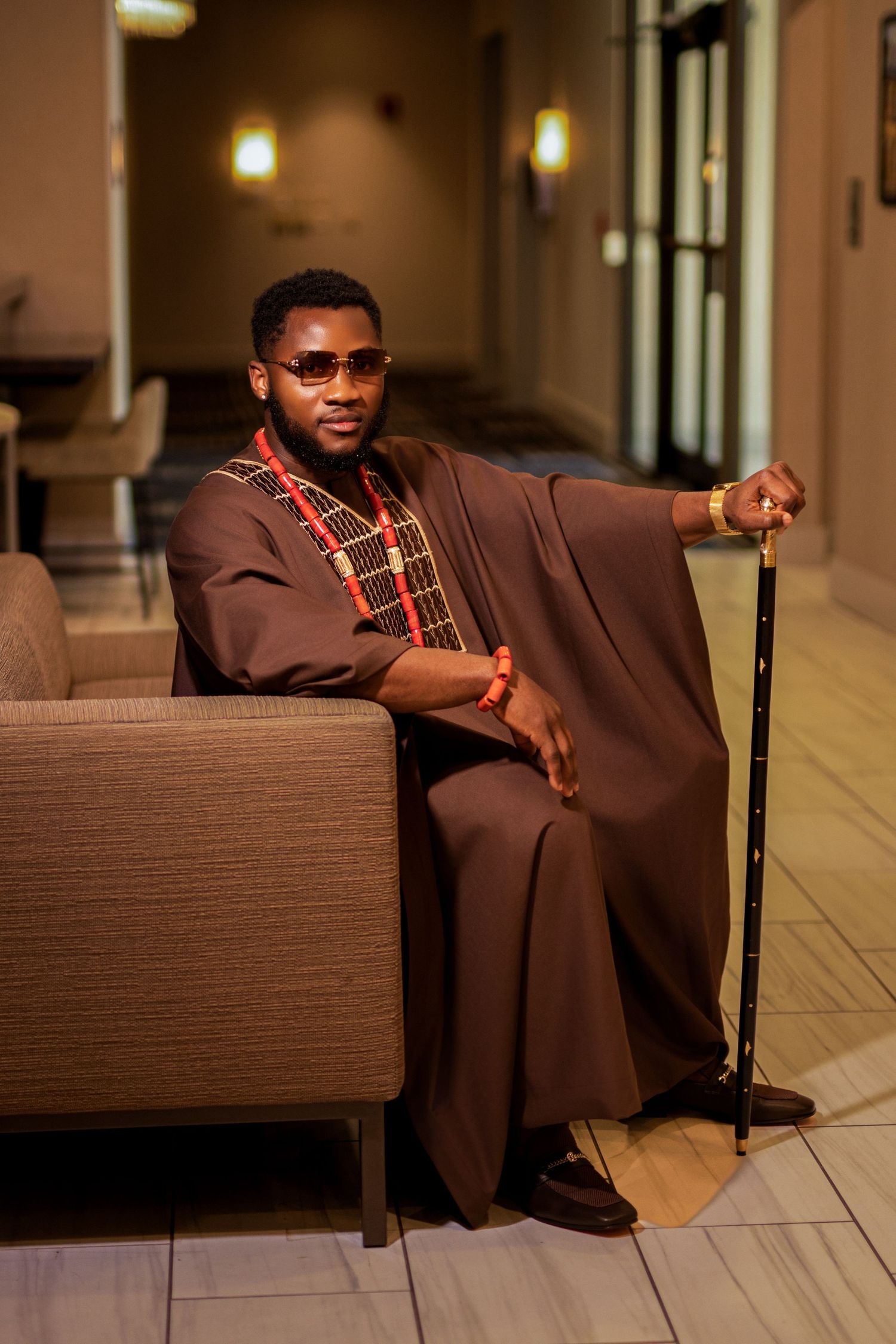Person in brown traditional attire relaxes on brown couch holding walking stick.