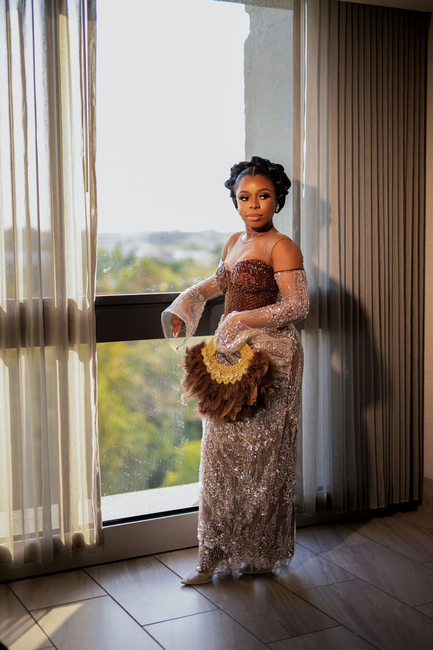 Person in sparkly brown gown with sheer sleeves poses by window holding decorative fan.