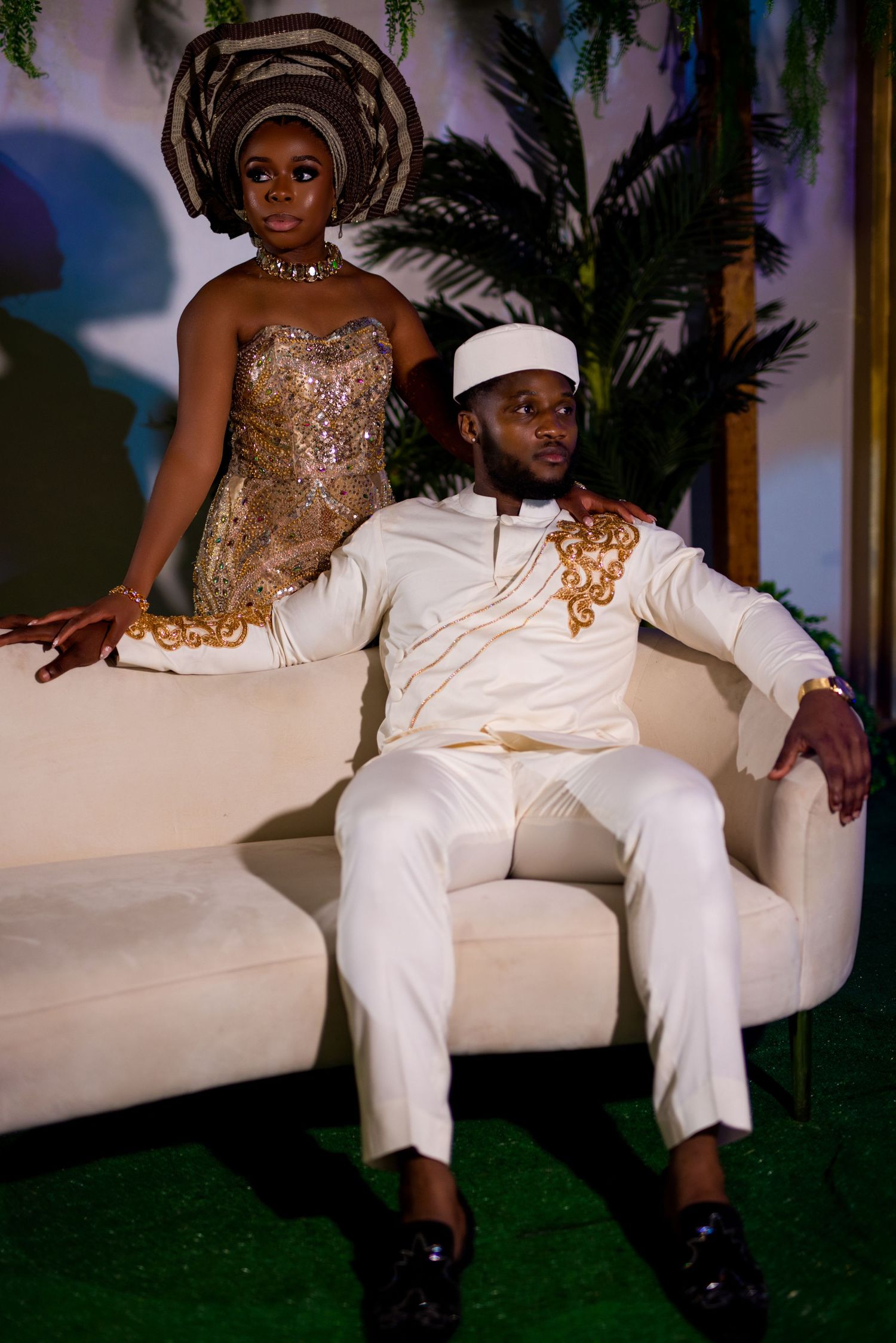 Couple in traditional African formal wear poses on white couch with tropical backdrop.