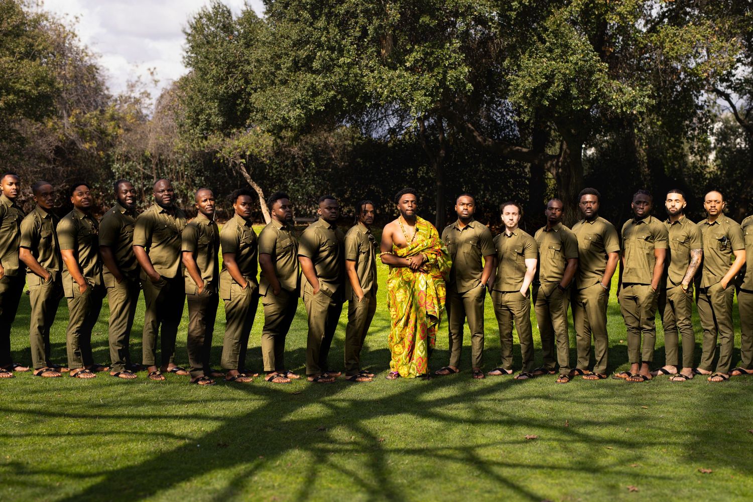 A group wearing yellow and khaki clothing stands in a line formation on a grassy field surrounded by trees.