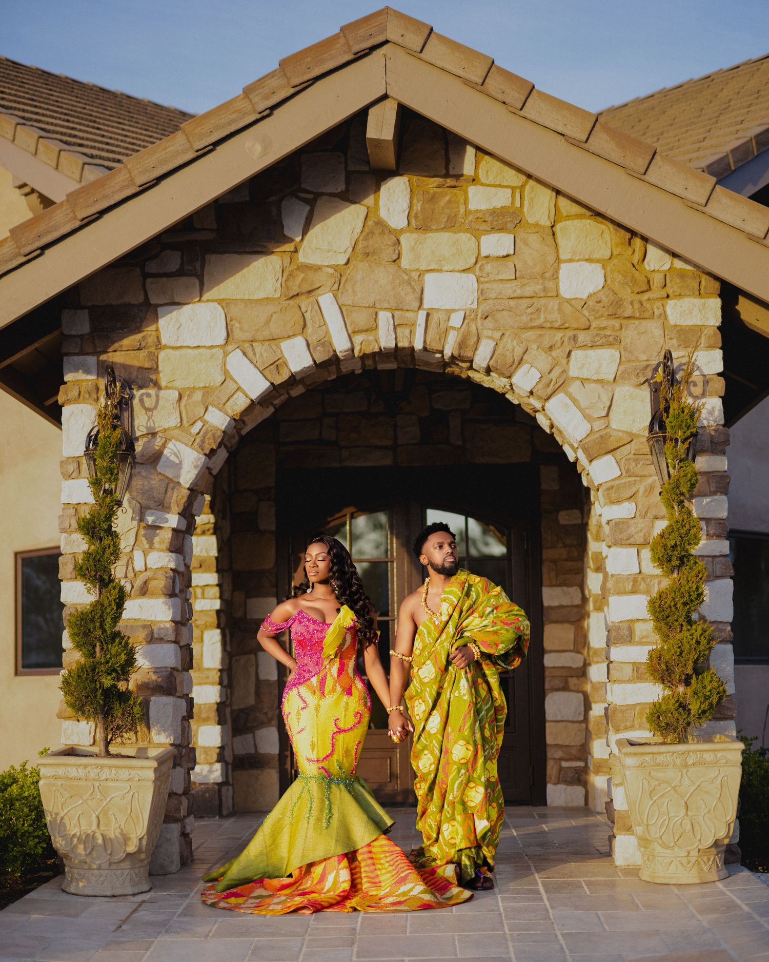 Two people in bright yellow traditional formal wear pose together under a stone archway entrance of a building.