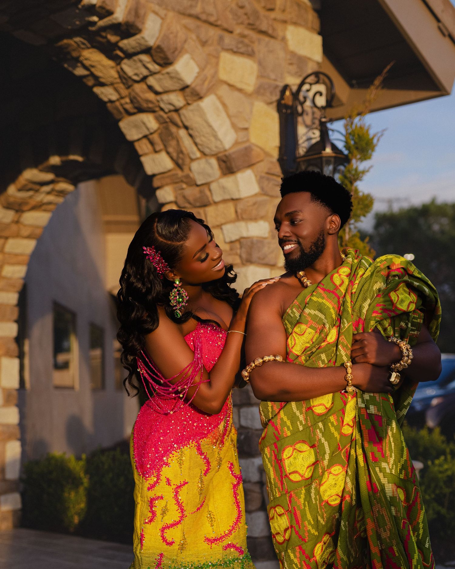 A couple wearing bright yellow and pink traditional African attire embrace lovingly in golden sunlight.
