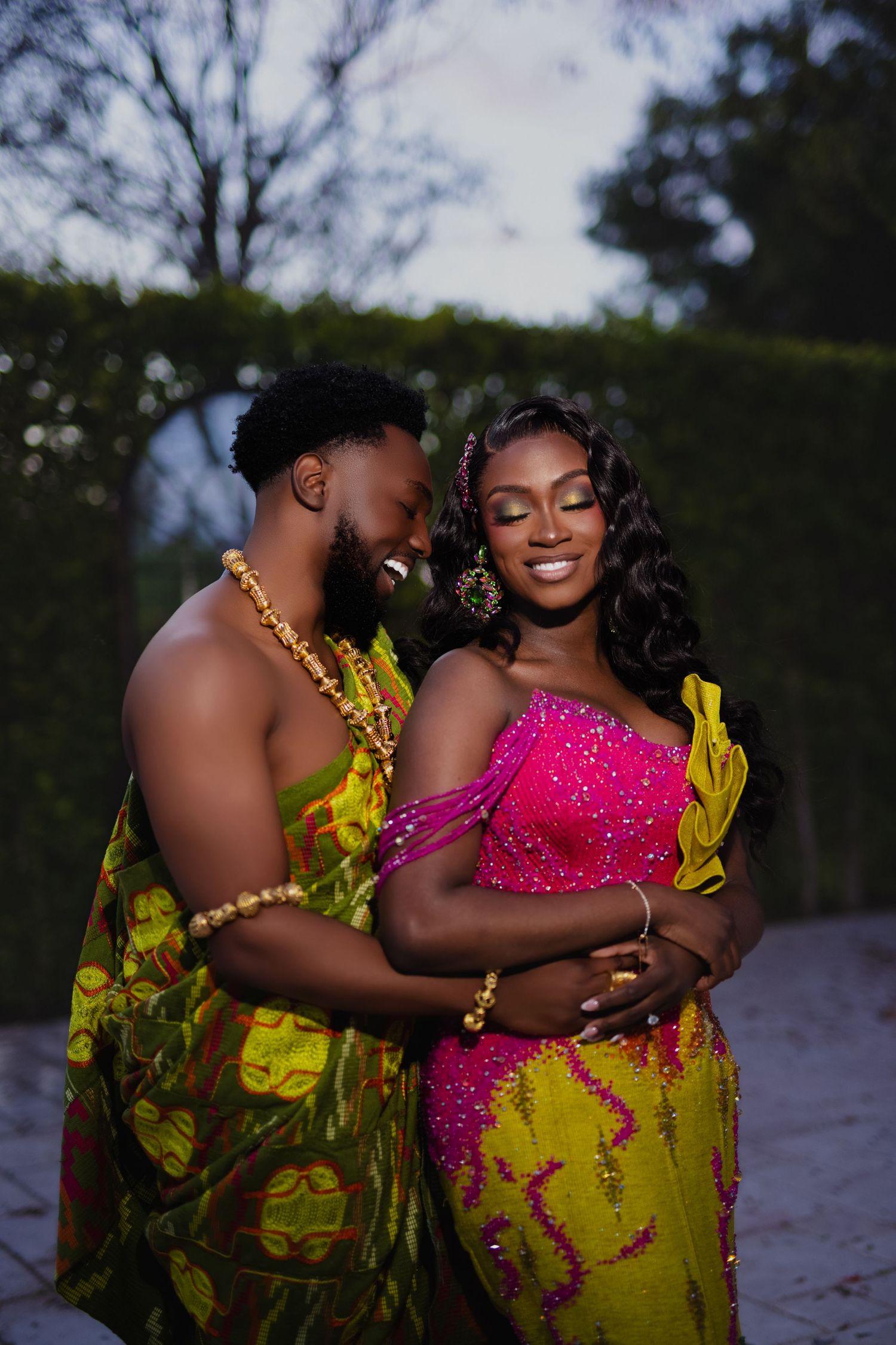 A romantic couple poses in vibrant yellow and pink traditional African-inspired formal wear during an outdoor photoshoot.