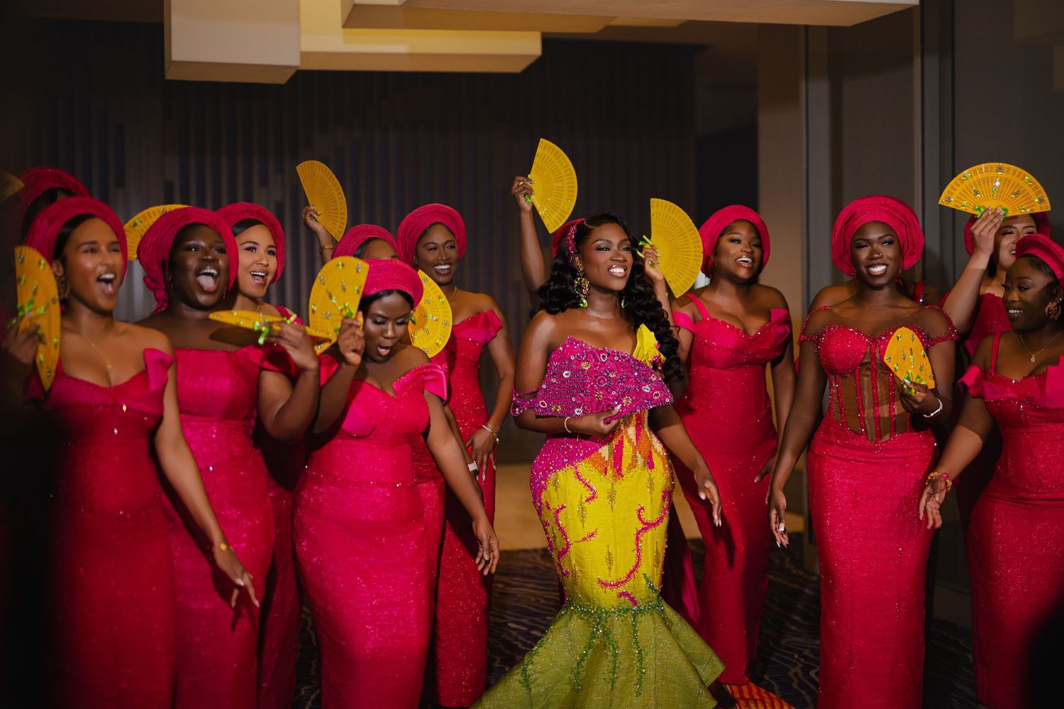Bridesmaids wearing vibrant red dresses and yellow fans pose with a bride in a yellow and pink mermaid gown.