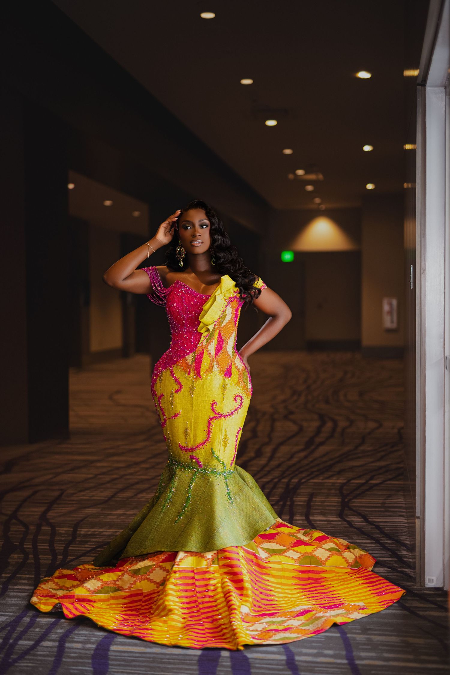 A vibrant mermaid-style gown in pink, yellow and green with dramatic ruffled train sits in an elegant hotel hallway.