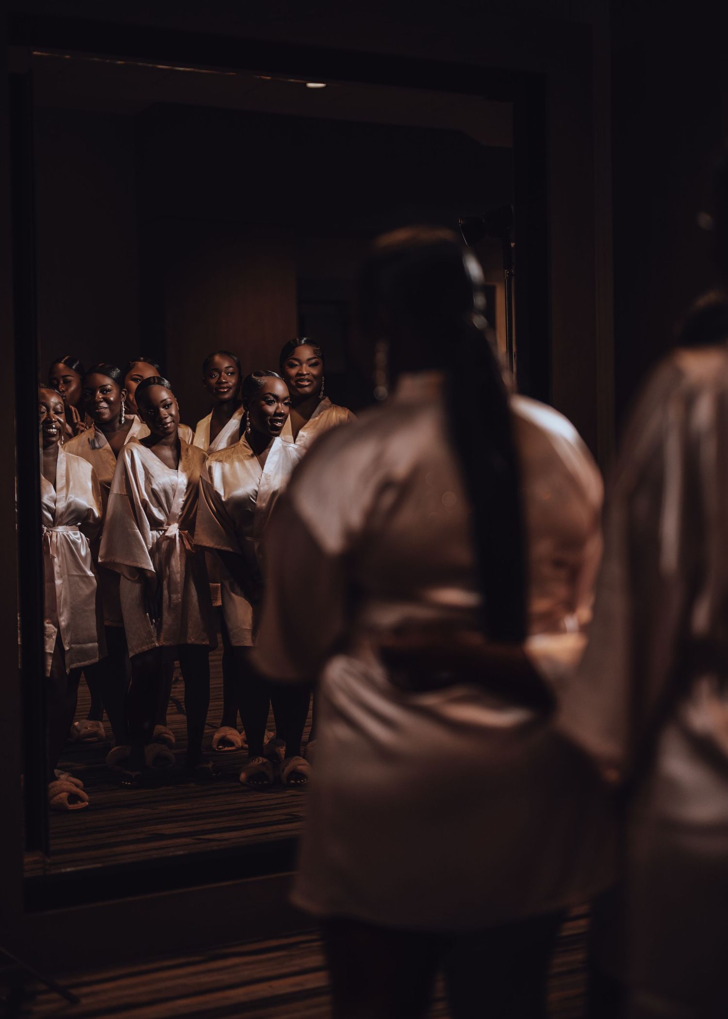 A group of wedding guests in white clothing gather and reflect in a dark mirrored hallway.