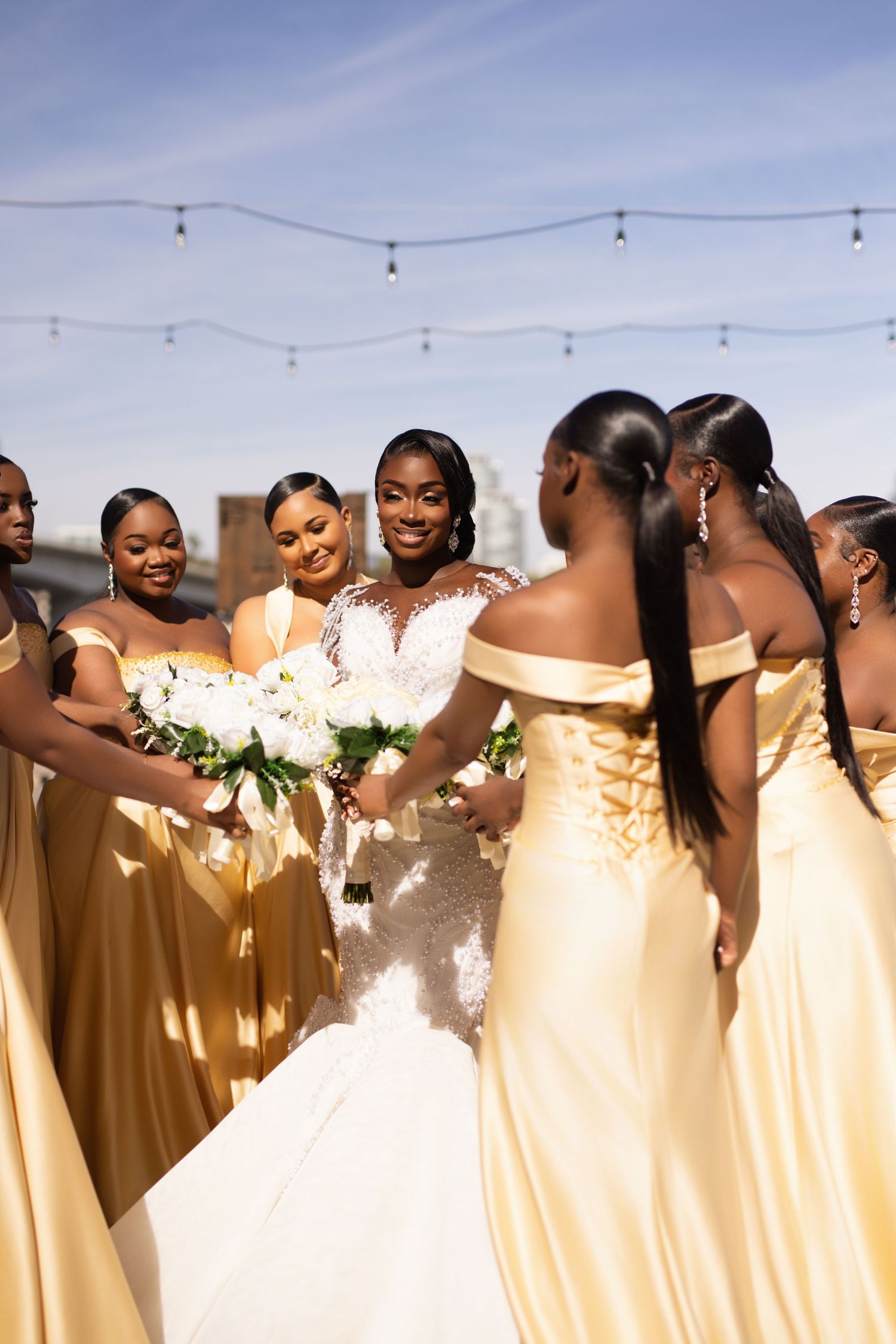 Wedding party in soft yellow bridesmaid dresses gather outdoors under string lights at sunset celebration.