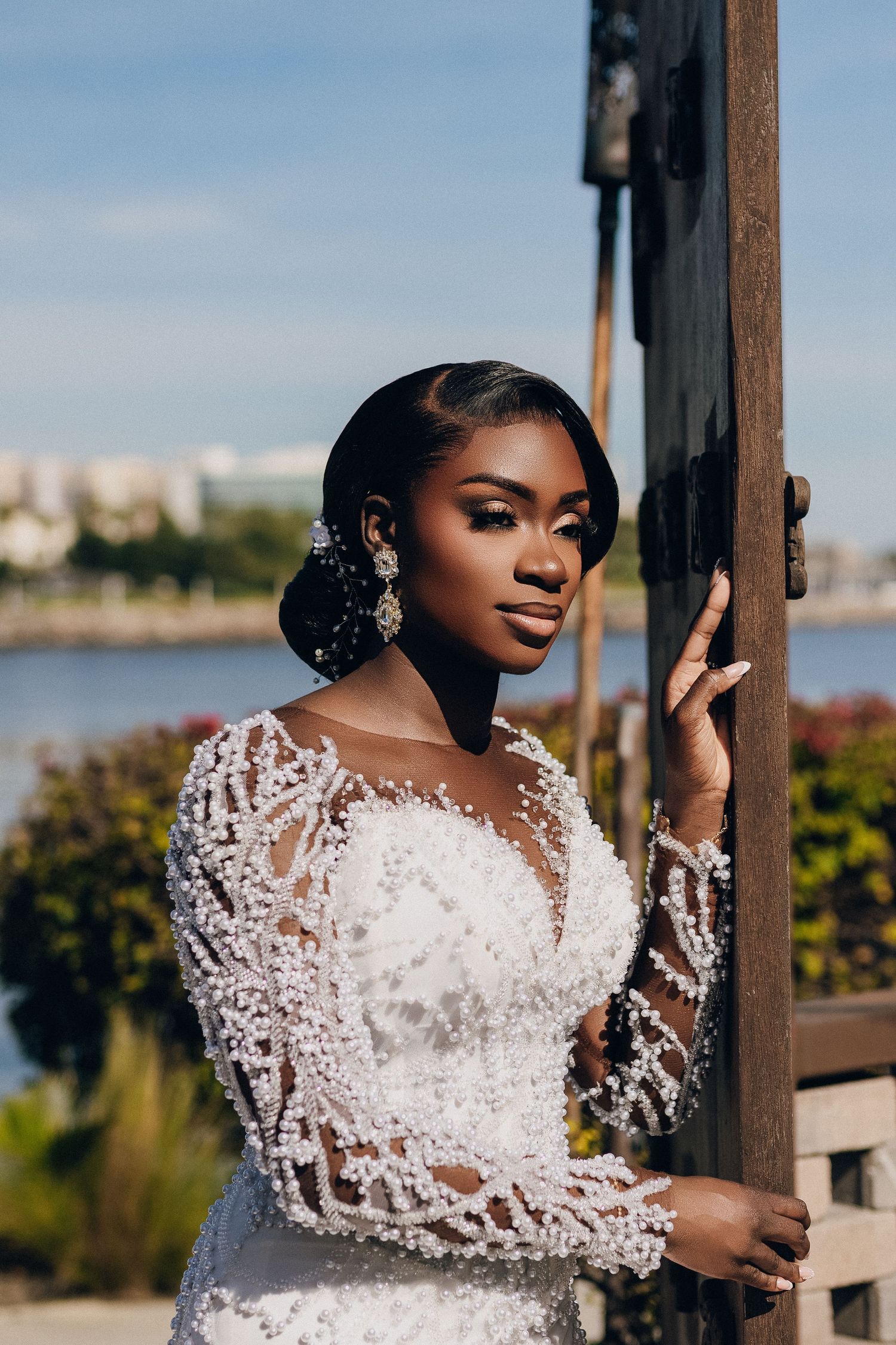 Elegant bride in a long-sleeved beaded lace wedding gown poses outdoors with a waterfront backdrop on a sunny day.