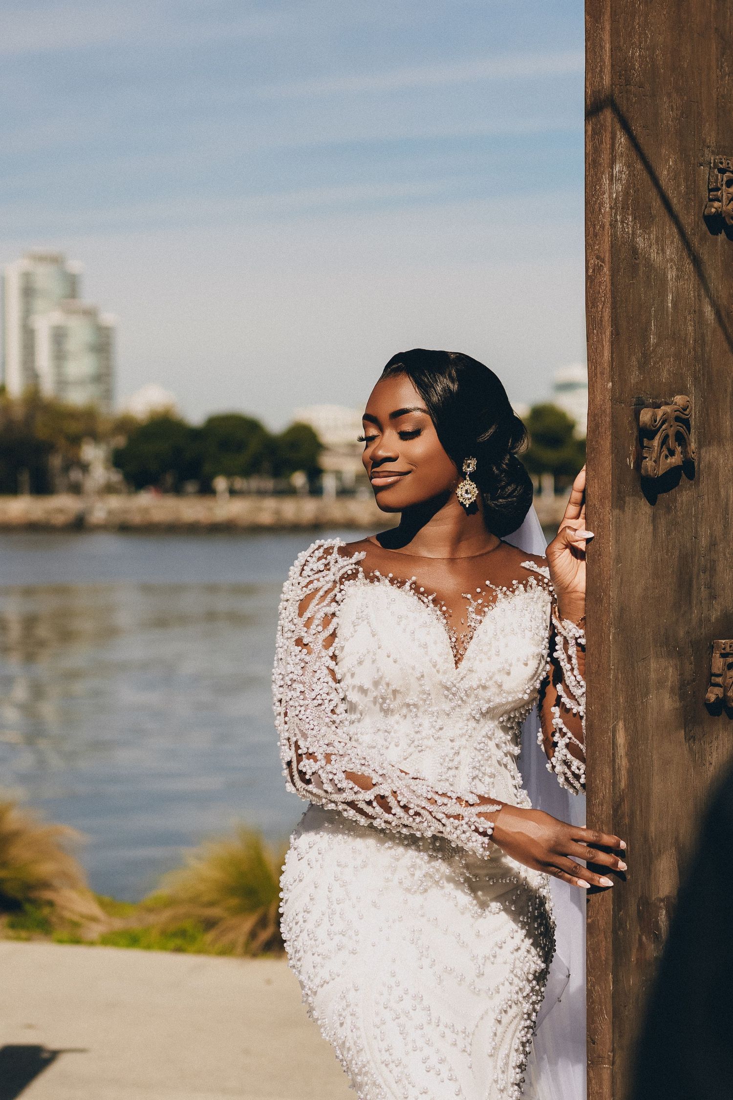 A bride in an elegant lace wedding dress poses against a wooden beam by the waterfront.