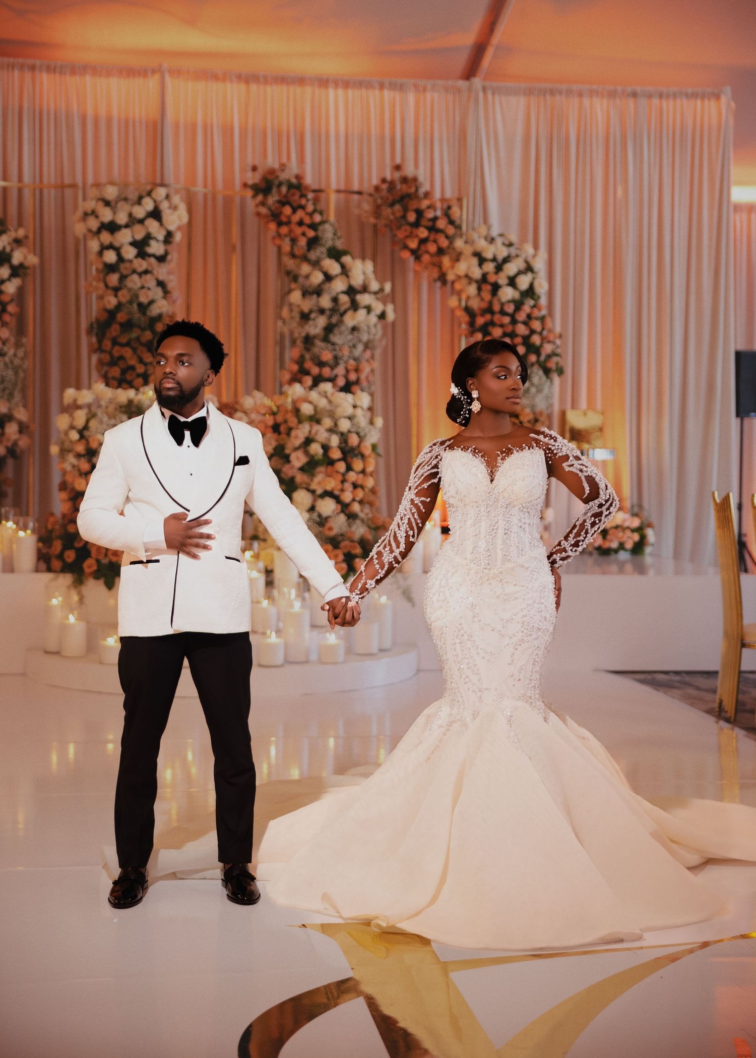 A couple in elegant wedding attire strikes a playful pose in front of a floral wall backdrop.