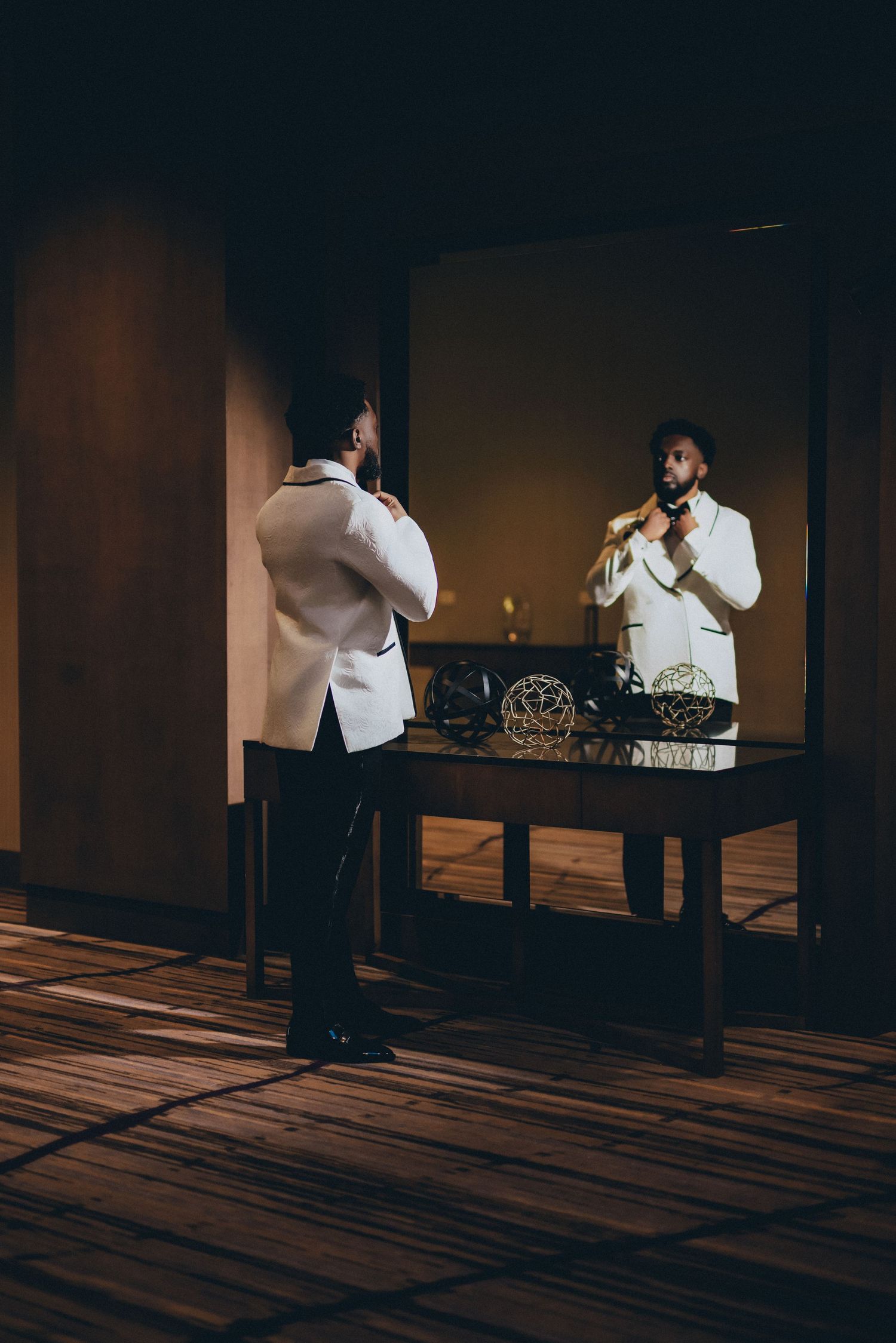 A person in a white suit adjusts their tie while looking in a mirror with dramatic moody lighting.