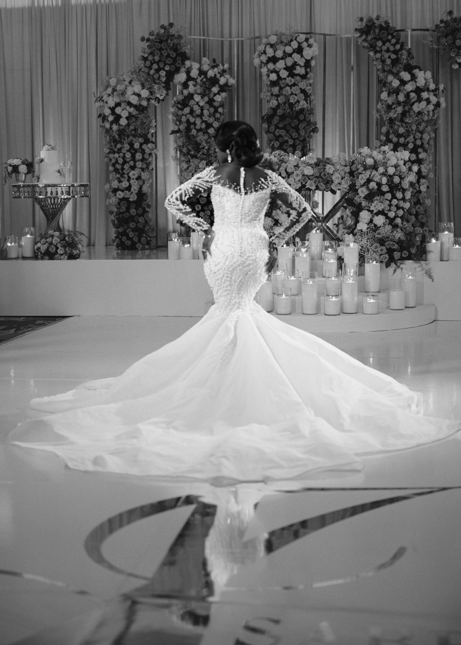 A stunning mermaid wedding dress with a dramatic train reflects in a mirrored floor amid floral arrangements.