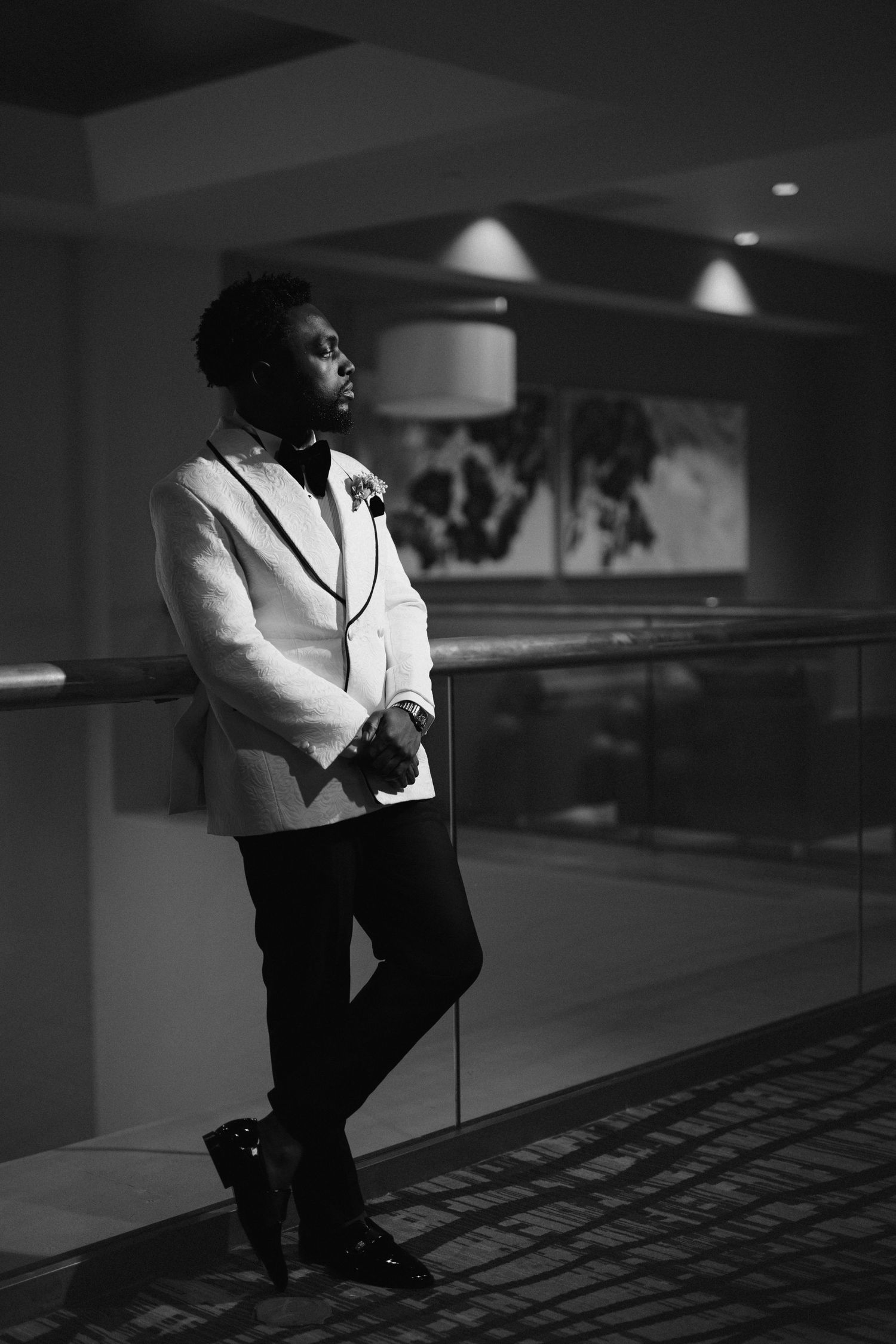 A person in formal black and white attire leans against a wall in a dimly lit hallway with modern lighting.