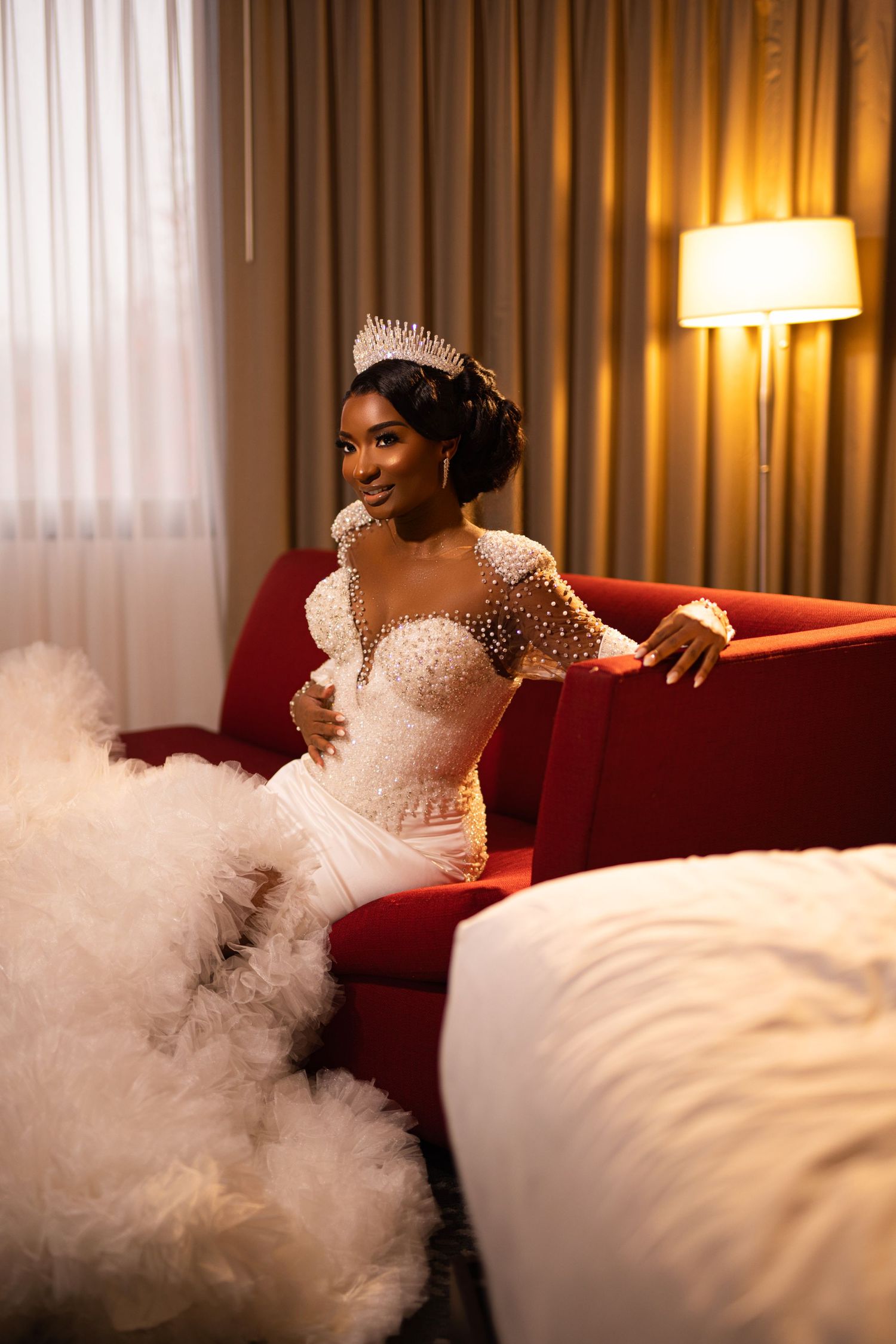 Bride in elegant lace gown poses on red couch with dramatic lighting and smoke effects.