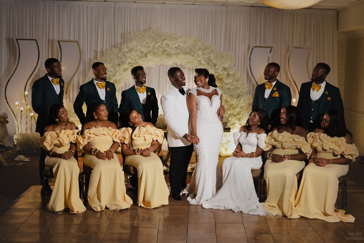 Wedding party poses together with bridesmaids in gold dresses and groomsmen in black tuxedos.