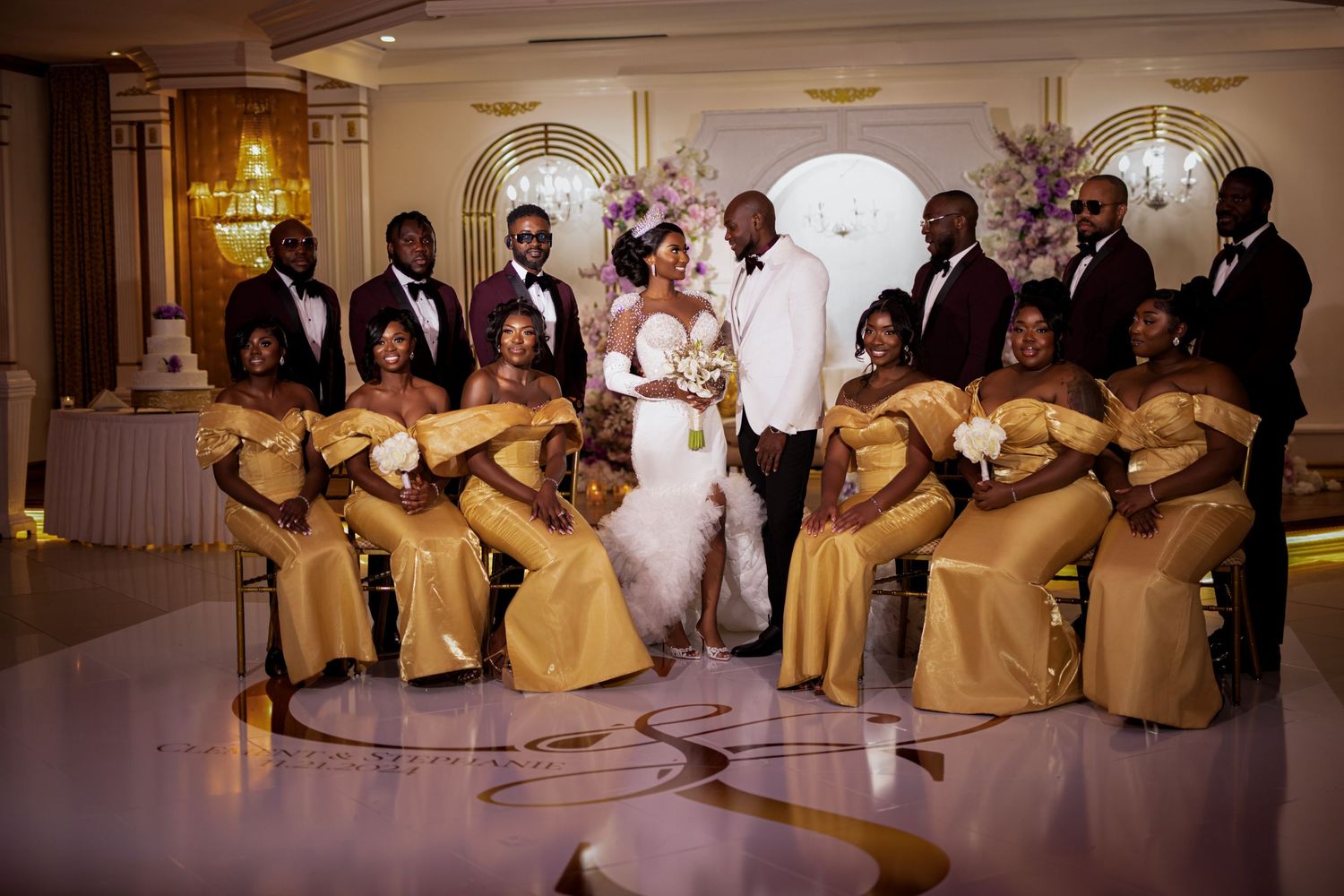 Wedding party poses in formal attire with bridesmaids wearing matching gold dresses in an opulent ballroom.