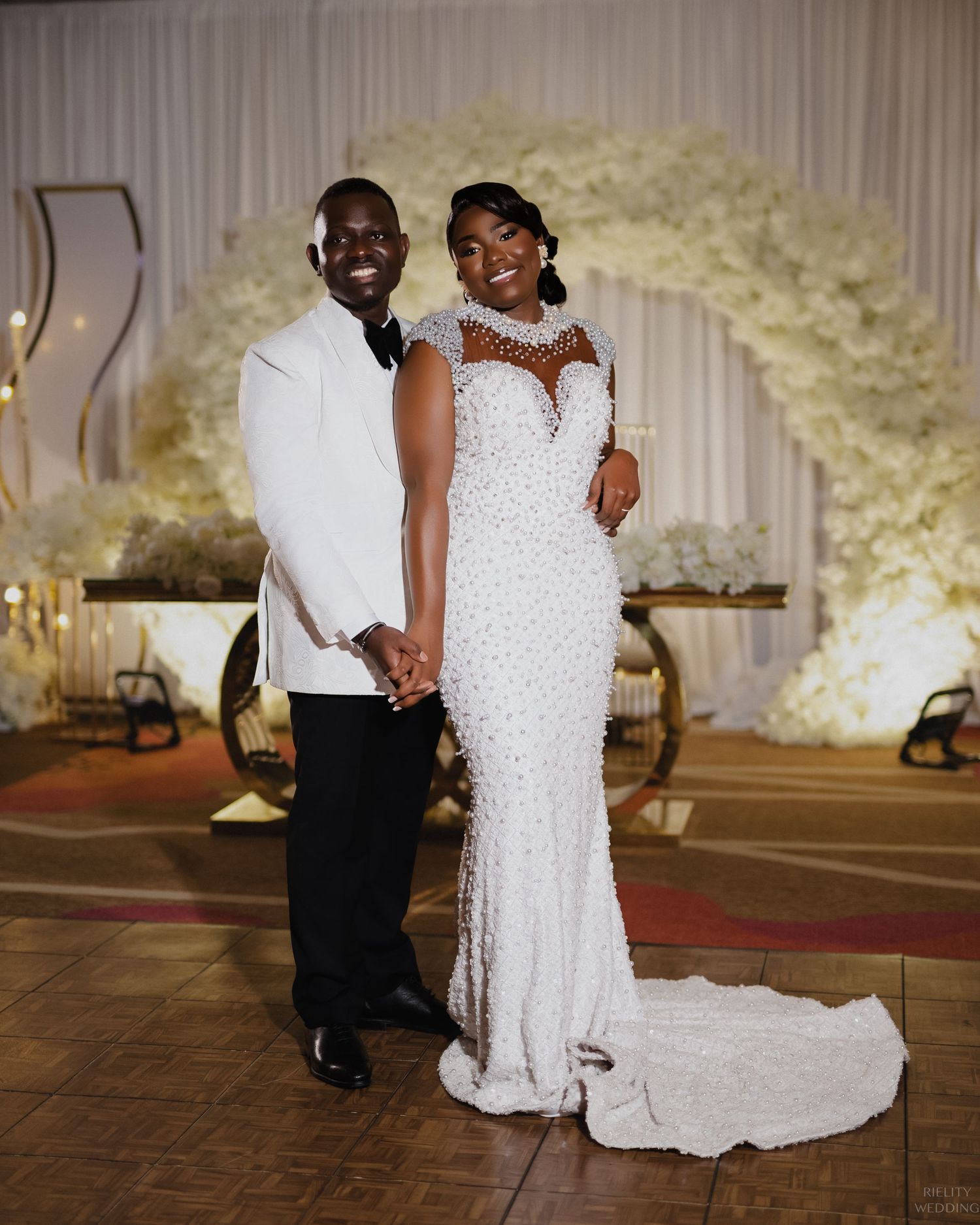 Couple in elegant white wedding attire pose under floral arch at reception venue.