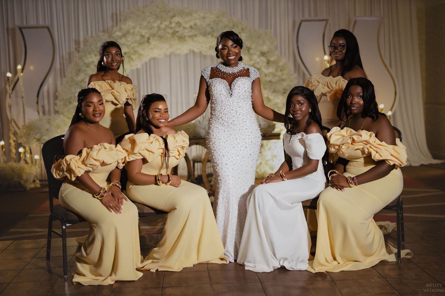 Wedding party in white and yellow dresses pose together under floral arch.