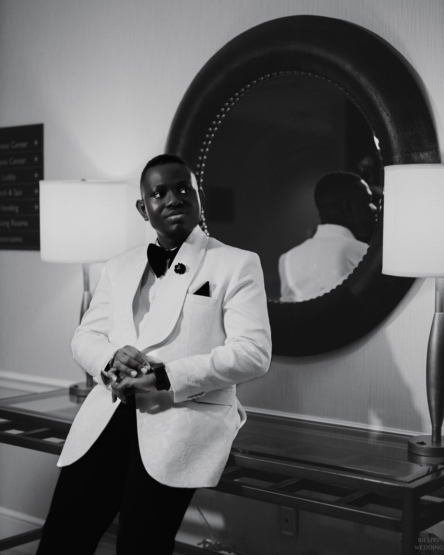 Groom in white tuxedo jacket and bow tie poses near decorative round mirror.