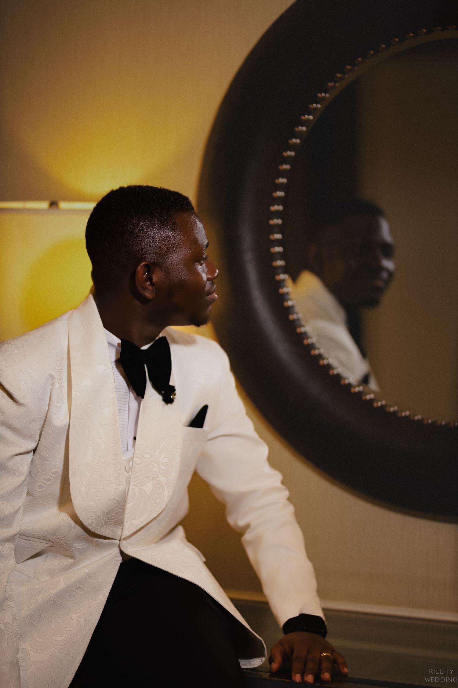 Side profile of groom in white tuxedo jacket posing next to circular mirror.