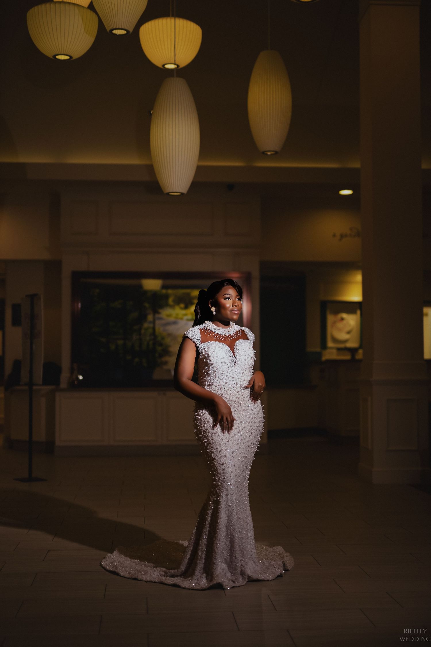 Bride poses in fitted beaded wedding gown under modern pendant lights in hotel lobby.