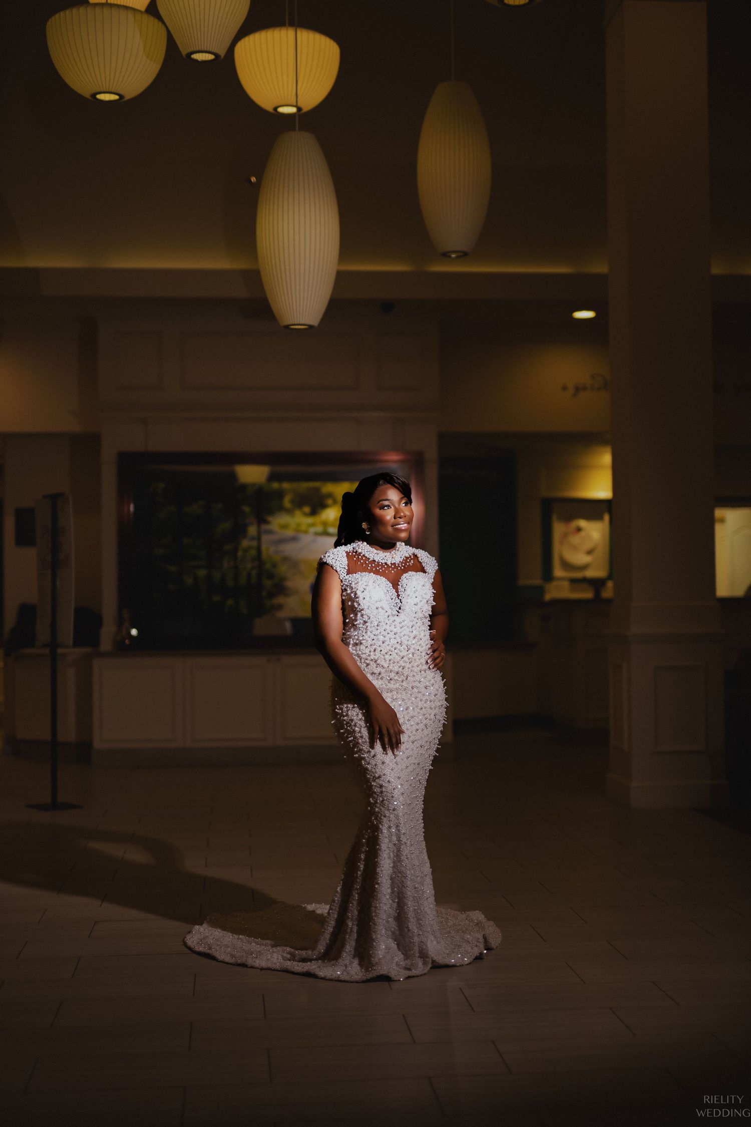 Elegant bride stands in fitted beaded gown under modern pendant lighting.