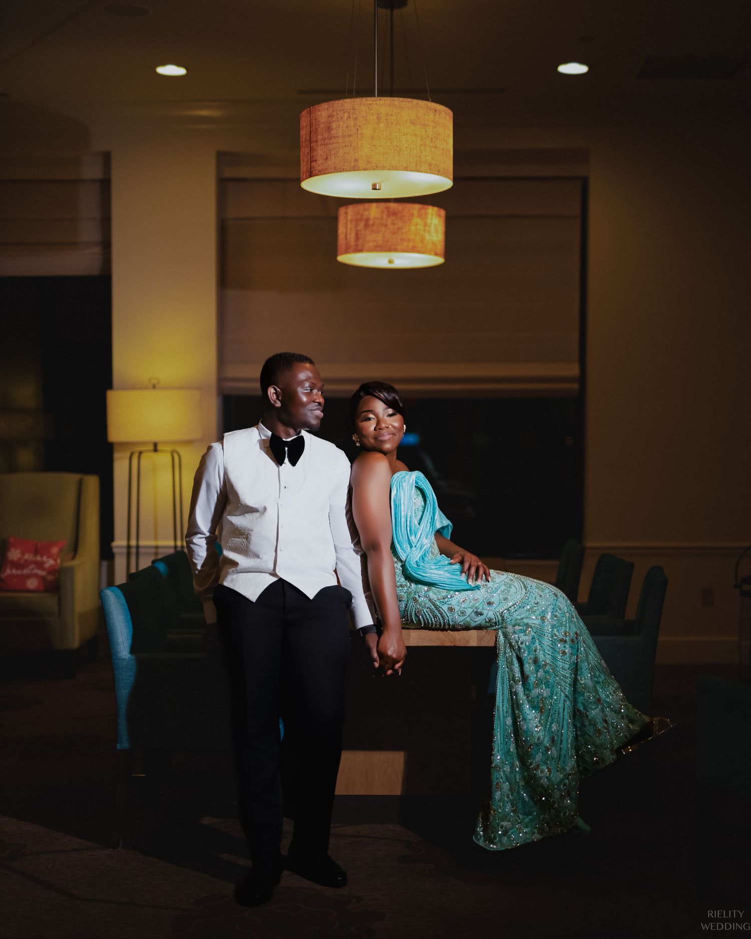 Couple poses together under pendant lights with groom in tuxedo and companion in teal gown.