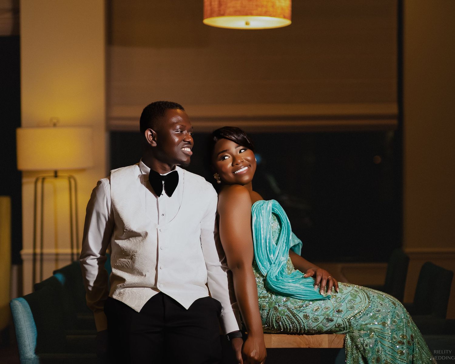 A couple in formal attire sit together in an intimate moment under warm lighting.