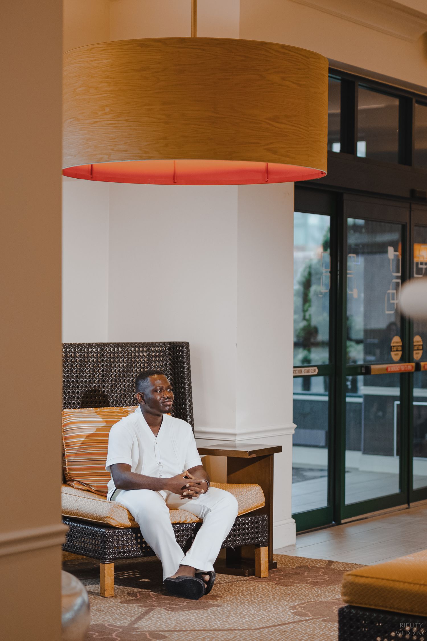 Person in white outfit sits on cushioned bench under large cylindrical pendant light.