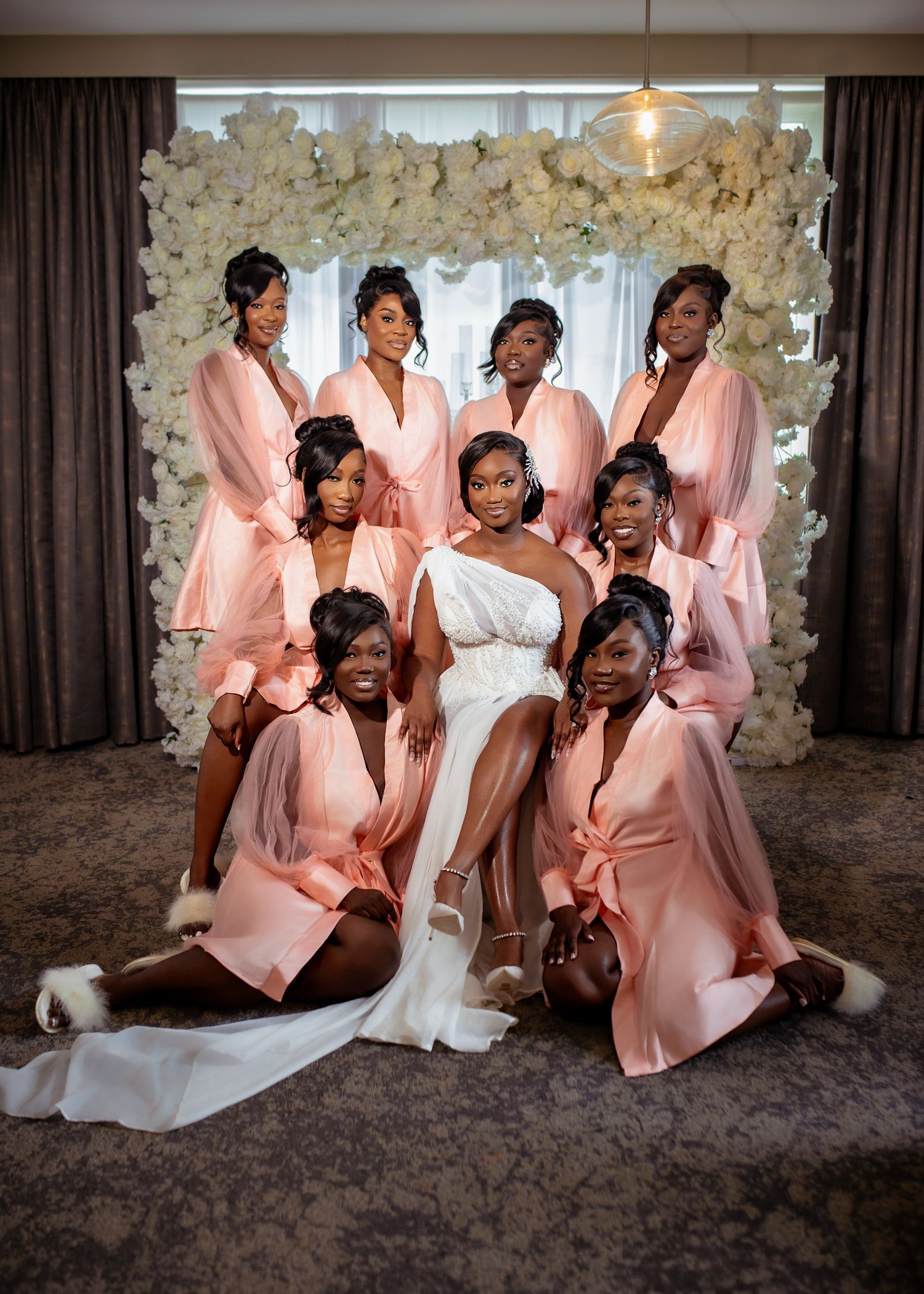 Wedding party wearing blush pink silk robes poses together on plush carpet with elegant floral backdrop.