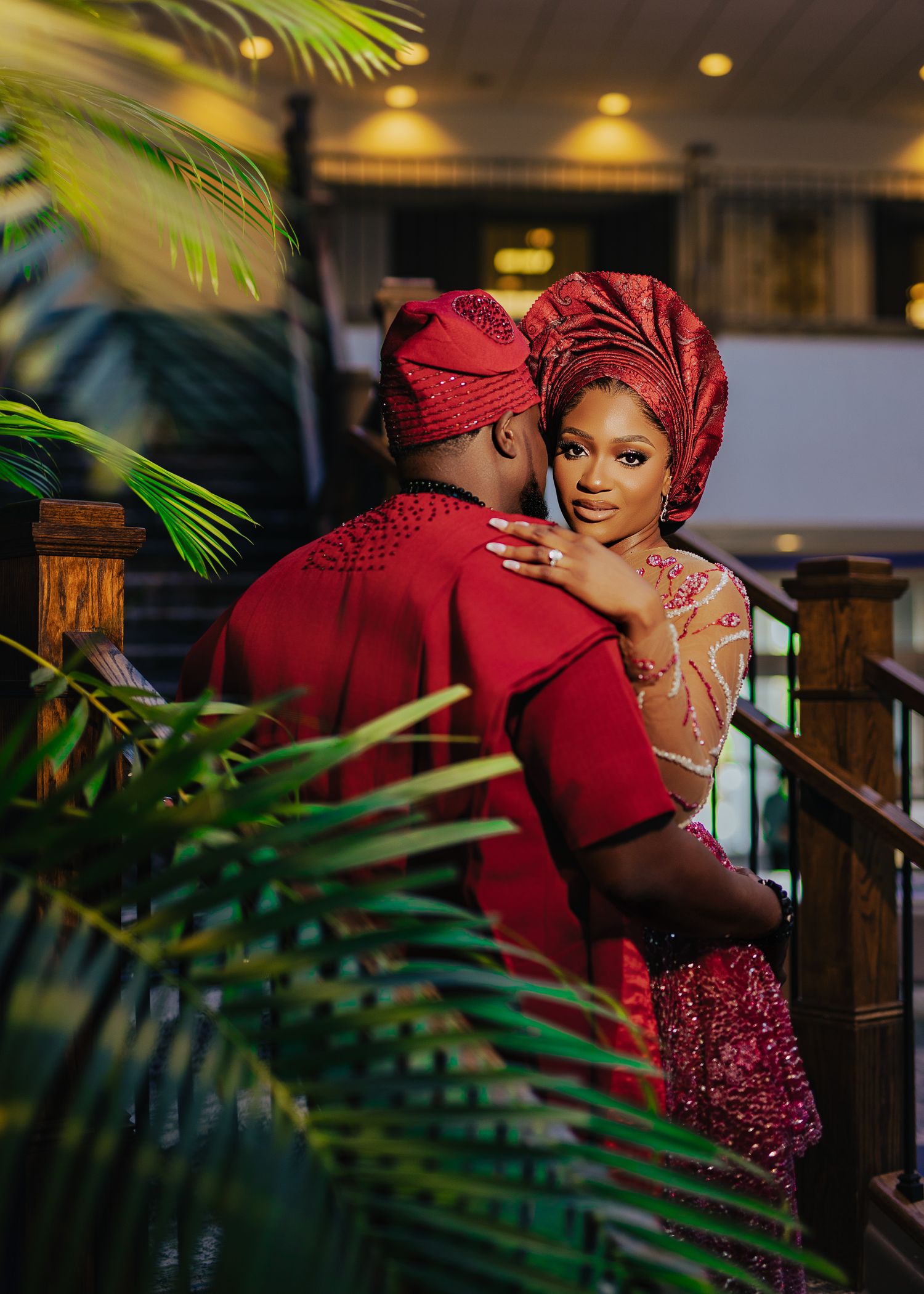 Couple in matching deep red traditional attire pose together among tropical palm leaves.
